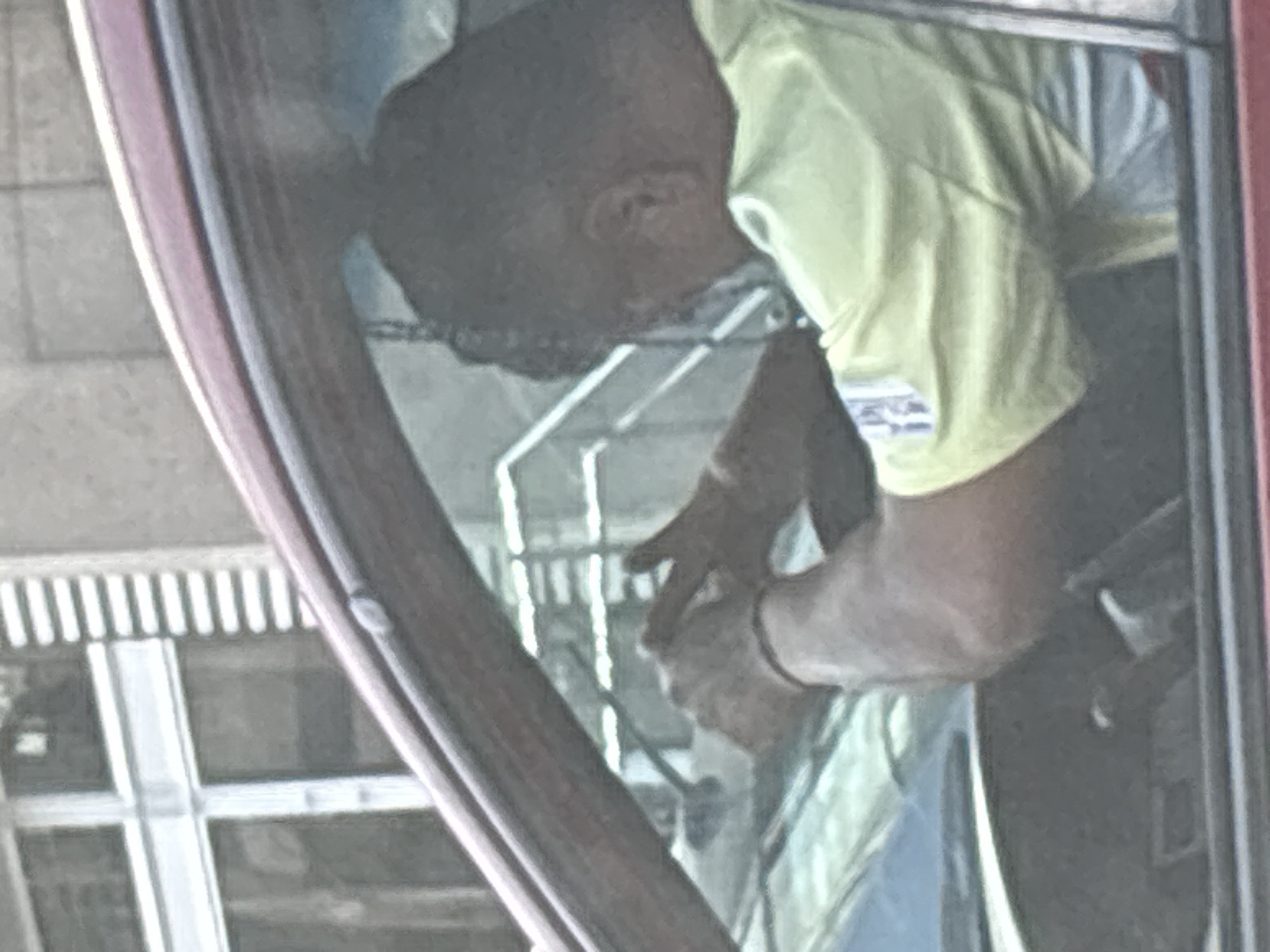 The image shows a person viewed through the slightly tinted side window of a vehicle, possibly a car or truck, with part of its reddish-brown frame visible on the left. The person has dark skin and hair, and is wearing a light-colored, short-sleeved shirt, possibly yellow or light green. Their head is bent downwards, indicating concentration on an activity.
The person's hands are visible and engaged in manipulating a small, light-colored object, which appears somewhat soft or fibrous. A dark band or bracelet is worn on their left wrist. The activity suggests a task requiring focused manual dexterity.
Through the vehicle's window, and as a reflection, a background resembling an urban environment is visible, characterized by vertical and horizontal lines, possibly from building windows, blinds, or a railing. The overall lighting suggests it is daytime. There are no discernible emotions or visible text in the image. The specified location context is Mexico City, Mexico.