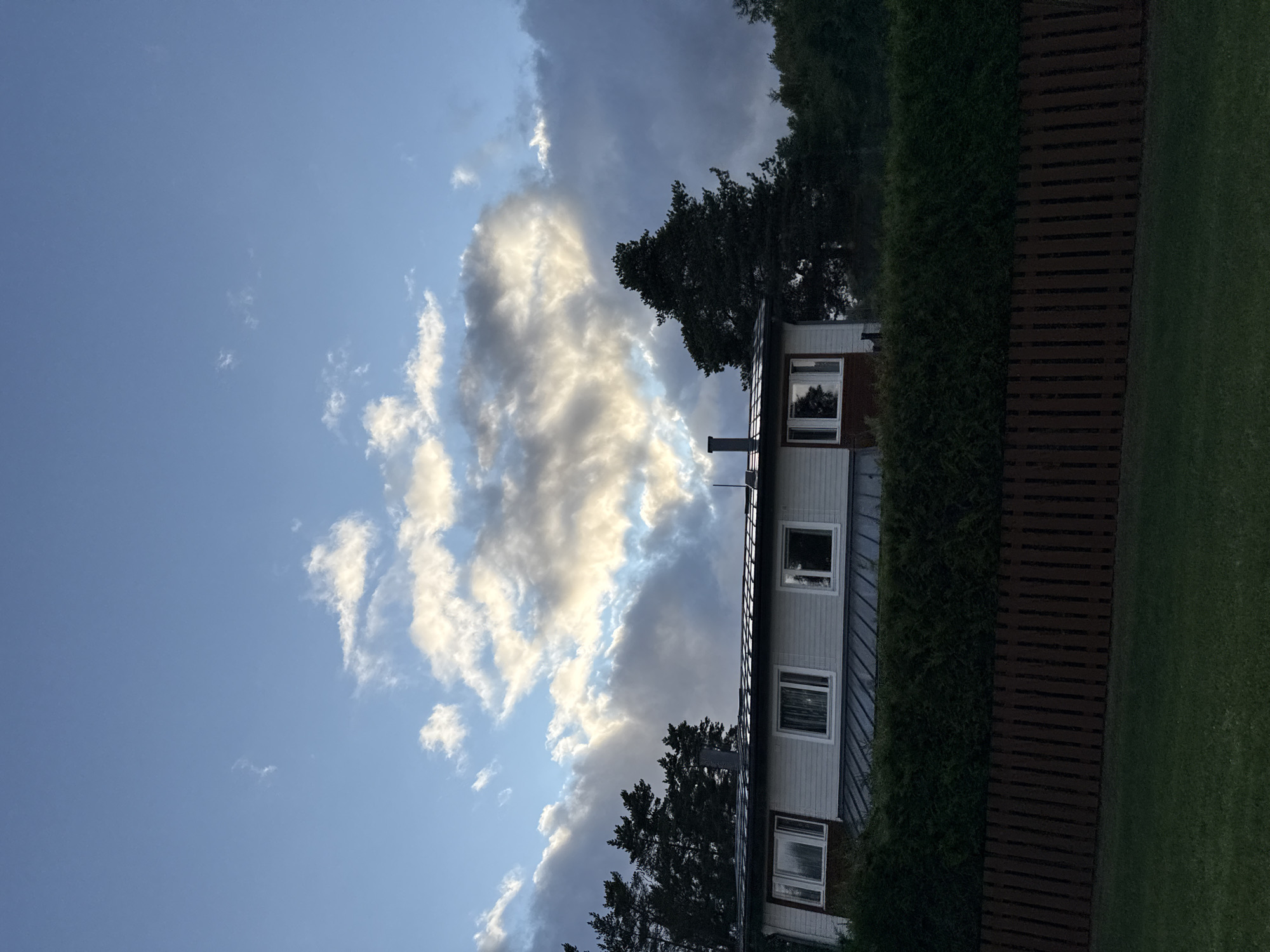 The image depicts a residential building with a light-colored exterior, featuring multiple windows, a dark-colored roof, and a chimney. The building is partially obscured by a dense green hedge and a dark wooden fence in the foreground. Behind the house, tall evergreen trees are visible against a sky with dramatic clouds, some of which are illuminated by sunlight. The scene appears to be set in Uuesalu, Estonia.