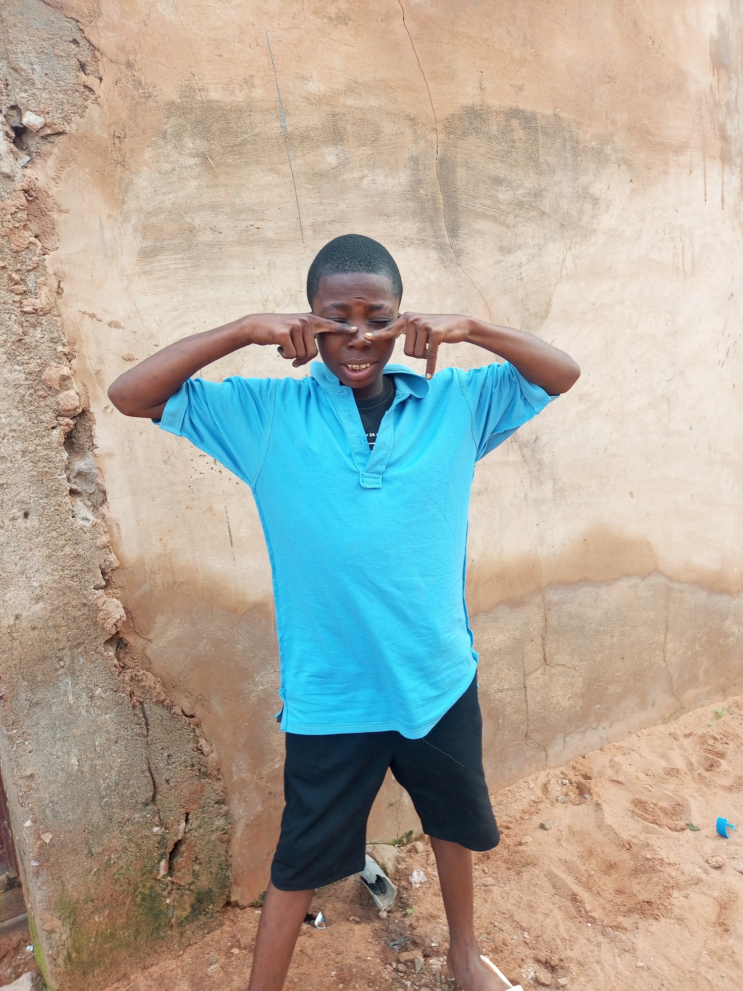 A young African male stands in front of a textured, pale brown, rough concrete wall. He is wearing a bright blue polo shirt with a black collar and black shorts. His right hand is raised, with his index finger touching his right eye, and his left hand is raised with his index finger touching his left eye, as if making a funny face. The background shows the wall, sandy ground, and some debris. The overall lighting suggests an outdoor daytime scene. The location is identified as Tempure, Nigeria.