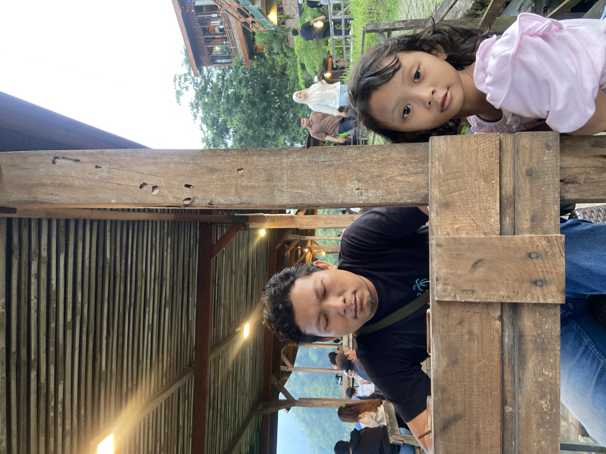 The image shows a man and a young girl posing, possibly for a photograph, in what appears to be an outdoor structure. The man, wearing a black t-shirt, is positioned with his face visible, though his eyes are closed or partially closed. Beside him, a young girl with curly hair is looking directly at the camera. She is wearing a pink top.
They are located within a wooden structure, likely a shelter or a viewing platform, with a corrugated metal roof visible above. The background shows a landscape with greenery, a building in the distance, and other people walking around. Given the location is Tamiajeng, Indonesia, it's possible the scene is within a recreational or tourist area.