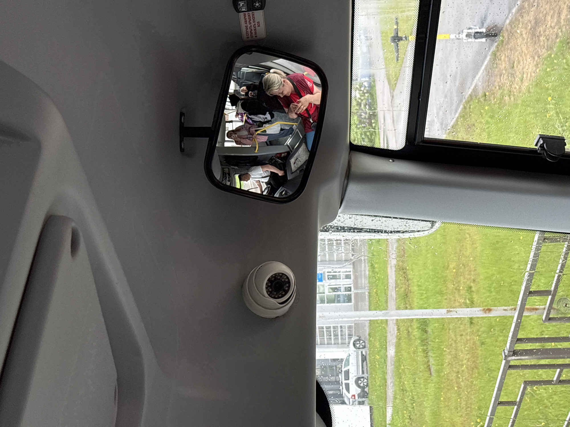 The image depicts the interior of a bus, likely a shuttle bus, possibly in Krasnoyarsk, Russia. The view is from the front of the bus looking toward the back, captured in a mirror above the driver. The reflection shows several passengers seated. A woman in a red shirt is visible near the front of the bus within the mirror. A white spherical security camera is mounted on the wall near the front window. Through the rain-spattered window, a view of what appears to be airport infrastructure and vehicles are visible. Specifically, a ladder, grassy areas, and a white vehicle are visible through the window, suggestive of an airport setting.