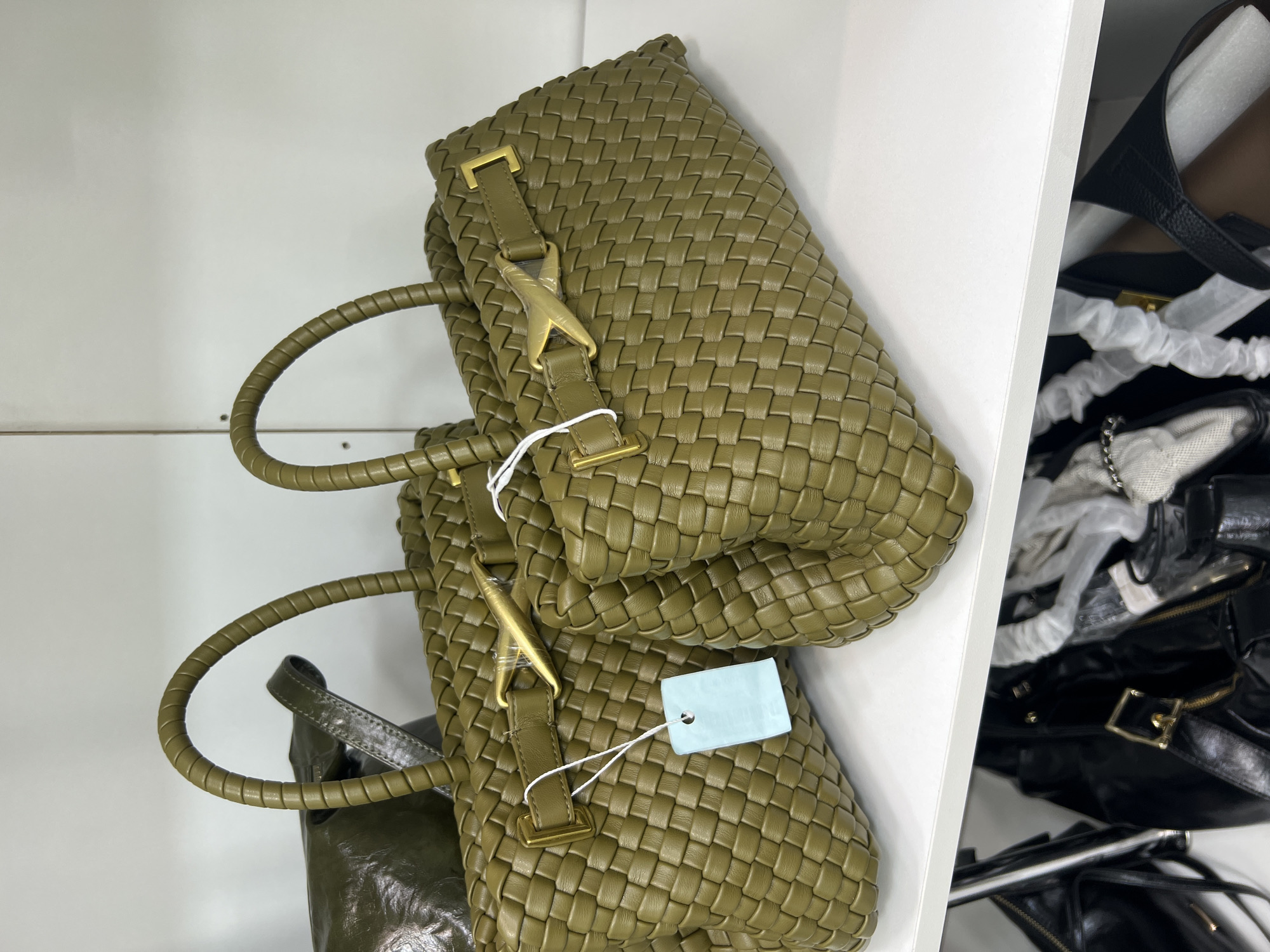 This image depicts two woven olive green handbags displayed on a white shelf. The handbags are positioned side-by-side, with the one on the right slightly closer to the viewer. Both bags feature braided handles and gold-toned hardware, including a distinctive clasp that resembles a 'X' shape. A light blue tag is attached to the left handbag by a white string. In the background and below the shelf, other dark-colored bags are partially visible. The overall environment suggests a retail or storage setting, likely a boutique or a closet. The image does not contain any elements that would allow for the identification of a specific location within Osh City, Kyrgyzstan.