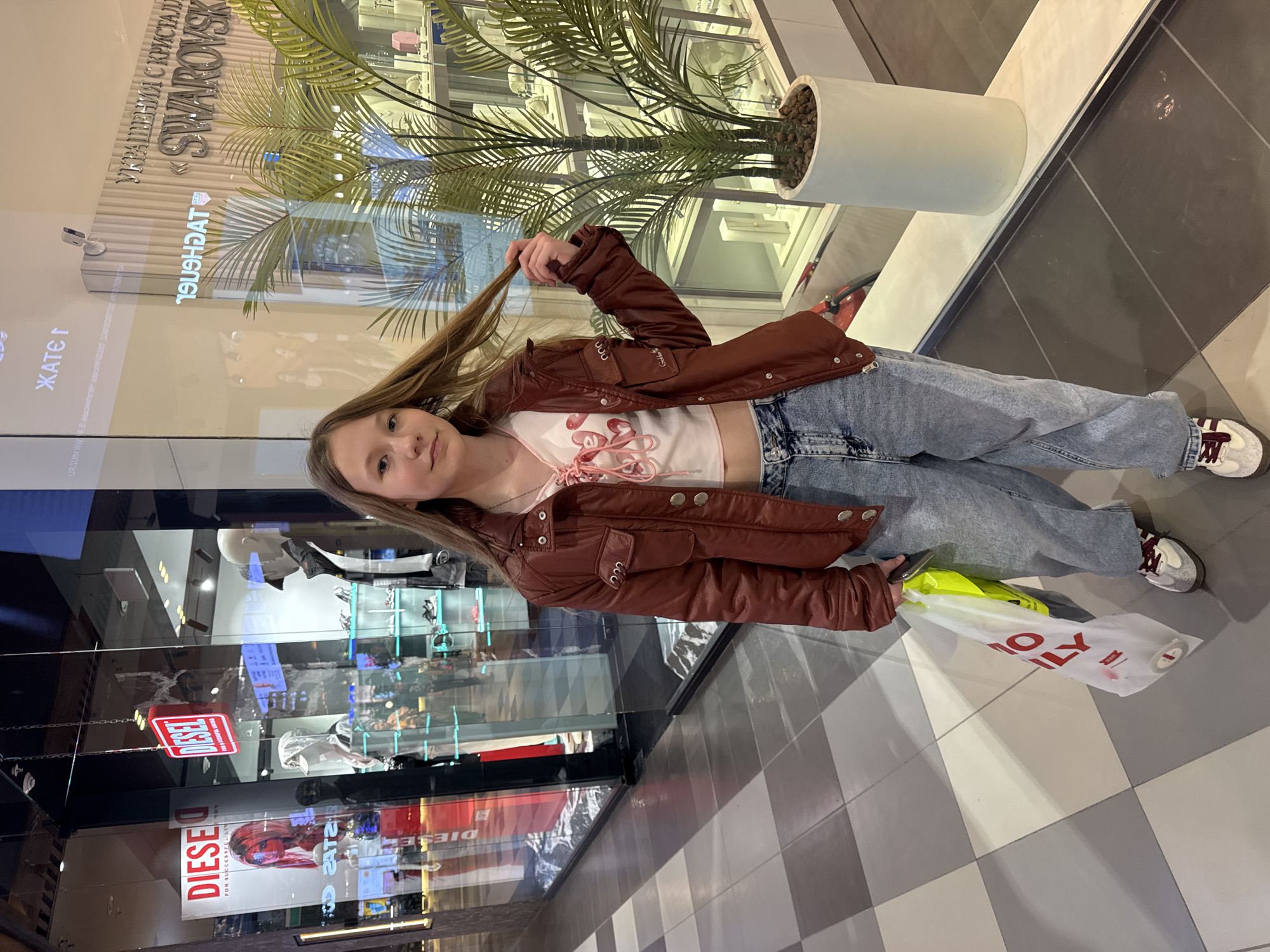 The image shows a young girl standing in what appears to be a shopping mall or retail environment. She is wearing a brown jacket, a white top, blue jeans, and sneakers. She is holding a strand of her hair in her hand and appears to be posing for the photo.
Behind her is a store window with various items on display, as well as a large sign that reads "SWAROVSKI." There is also a potted plant near the window. To the side, there is a sign with the text "DIESE" and "FOR SUCCESSFUL LIN" visible.
The flooring consists of gray and white square tiles. The scene is well-lit, suggesting an indoor environment.