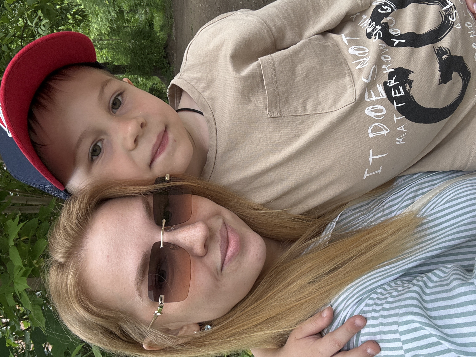 This image, taken in Rusanovo, Russia, shows a woman and a young boy together outdoors, surrounded by green foliage. The image is rotated 90 degrees counter-clockwise.
In the foreground, a woman with long blonde hair wears rimless sunglasses with amber lenses and a light-colored top featuring thin white and light green stripes. She has freckles on her nose and cheeks and a relaxed, slightly smiling expression. Her hand is visible resting on her striped top.
Behind and slightly above her, a young boy, appearing to be 4-7 years old, wears a red and navy baseball cap and a light brown long-sleeved t-shirt. He looks directly at the camera with a gentle smile. A black choker is visible around his neck.
The background consists of blurred green trees and leaves, suggesting a park or garden setting, with a faint dirt path visible. The lighting indicates bright daytime conditions.
On the boy's t-shirt, there is prominent black and white text. Large stylized black shapes resemble a '33' or similar graphic. White text spells out the phrase: "IT DOESN'T MATTER HOW STRONG YOU ARE". The image conveys a casual and warm moment between the two.