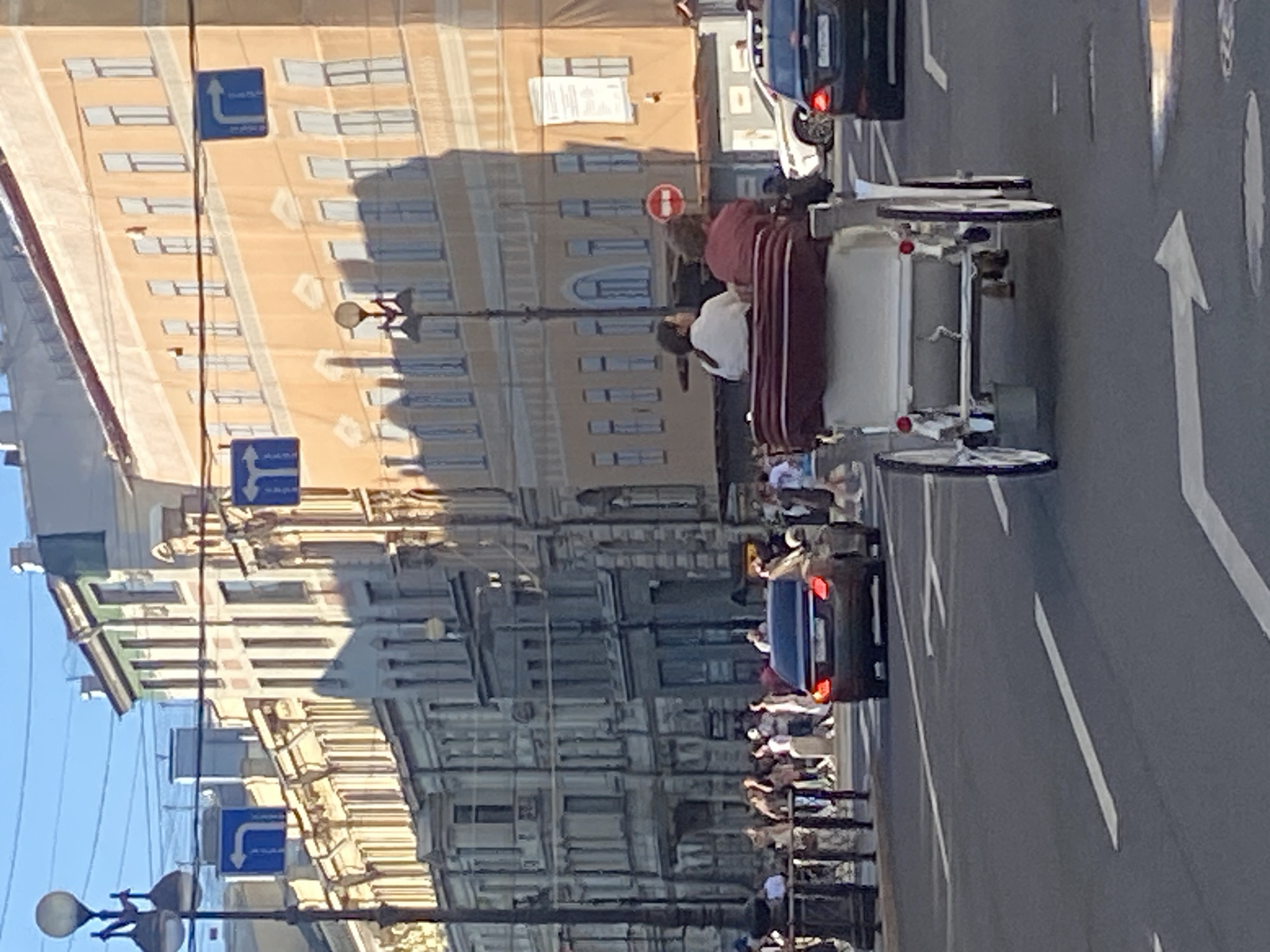The image depicts a street scene in Saint Petersburg, Russia, likely during daylight hours. In the foreground, a white horse-drawn carriage with two passengers is moving along the road. The passengers are seated facing forward, with one individual wearing a white shirt and the other a maroon shirt. The carriage has a maroon canopy and a white body.
The road is asphalt with white lane markings, including an arrow pointing left. Several cars are visible on the road. A dark sedan with its taillights illuminated is in the right lane ahead of the carriage, and a white SUV is to its right.
The background features buildings lining the street, characteristic of Saint Petersburg's architecture. The buildings are multi-story structures with ornate facades, some painted in light colors and others in a pale orange hue. Pedestrian traffic is visible on the sidewalks, with a group of people gathered near a dark railing in the mid-ground. Streetlights and traffic signs, including blue signs with white arrows indicating direction, are also present. The sky is clear and blue, suggesting good weather. The overall impression is a busy urban environment with a mix of modern vehicles and traditional horse-drawn transport.