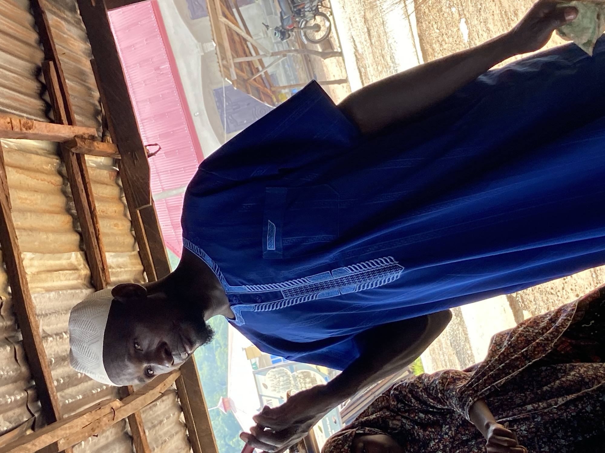 A man in a white kufi and a blue kaftan stands under a corrugated metal roof in Mararaba, Nigeria. He is holding a red object in his left hand, possibly a phone. A woman in a patterned headscarf and dress is partially visible to his left. In the background, a motorcycle is parked under a wooden shelter. The scene appears to be an outdoor market or communal area.