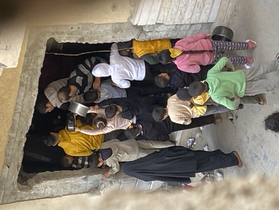 The image captures a crowded scene at the entrance of a concrete structure in Khan Yunis, Palestinian Territory. A large group, predominantly children and young adults, are densely gathered at and within a rough, unfinished doorway. Several individuals, including a young person in a light pink top centrally positioned, are holding silver-colored metal pots or bowls, many outstretched upwards into the dark interior of the building, indicating a distribution or collection activity. An adult male in a yellow hoodie and another in a striped shirt are visible within the doorway, interacting with the crowd. To the left, an adult female in a black niqab and dark clothing stands partially obscured. Additional children are visible in the foreground and middle ground, some looking towards the activity at the entrance. One girl in a pink top with braided hair holds a black bucket at her side. The ground outside the entrance is a light-colored paved surface.