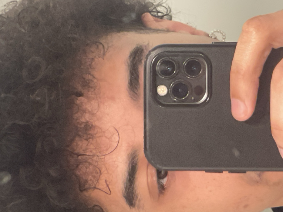 A close-up self-portrait image of an individual is depicted in Montigny-le-Bretonneux, France. The frame captures the upper portion of the subject's head and face, from the dark, curly hair at the top to the approximate mid-nose level. Dark, arched eyebrows are visible above dark brown eyes, with the left eye (viewer's left) being more fully present in the composition. The subject exhibits a light brown complexion. A black smartphone, featuring a rear triple-lens camera system and a flash module, is held centrally by the subject's right hand. The device is enclosed within a matte black, textured protective case. This phone partially obstructs the lower half of the subject's face. A small, circular earring, appearing to be composed of multiple small beads or pearls, is visible in the subject's right earlobe. Faint stubble is discernible on the left side of the subject's upper lip/chin area. The background is a plain, uniformly lit, light-colored surface, consistent with an interior wall.