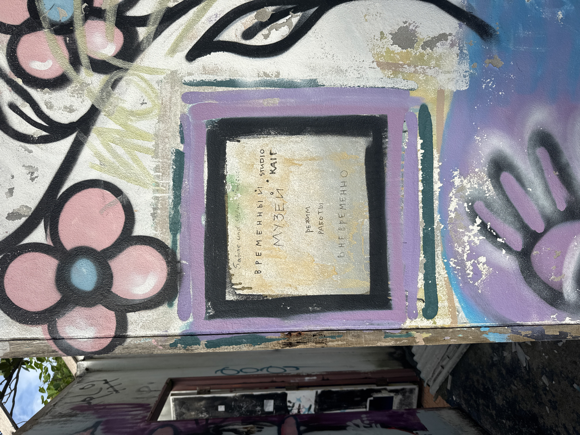 The image shows a section of graffitied wall, likely an exterior surface. There's a large abstract floral design with pink petals outlined in black. Above and to the right is a stylized eye. At the center is a square frame painted in black and lavender, containing text in what appears to be Russian. The text includes the words "МУЗЕИ" (Museums) and possibly information about hours of operation or a studio. The wall surface is weathered and peeling. Below the graffitied section is a lower surface, possibly a roof or ledge, with additional graffiti. There is no verifiable information that the image can be confirmed to be from Baan Thong Nang, Thailand.