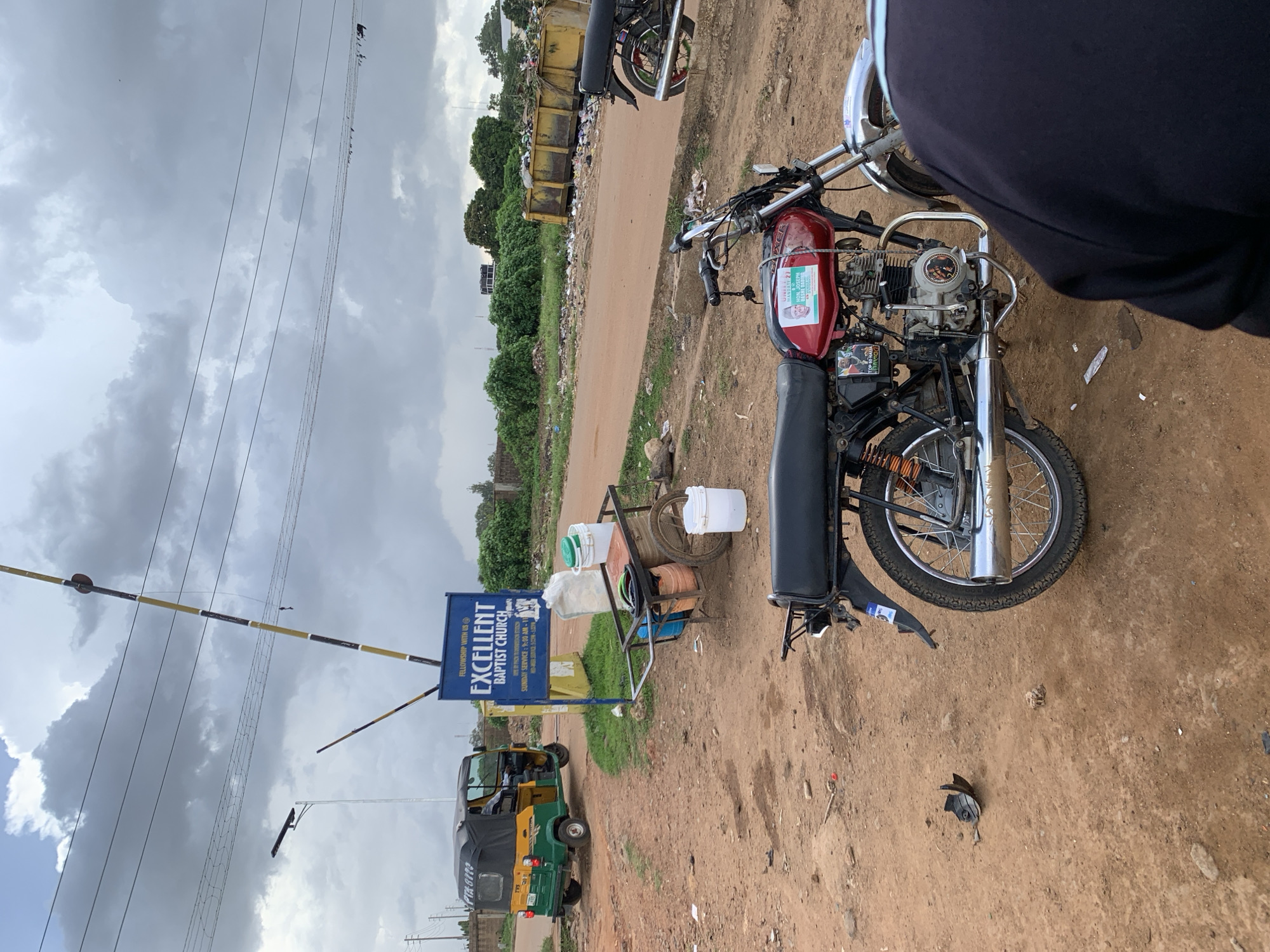 An outdoor, eye-level, medium shot captures a dirt road and its surroundings in Barakin Lerawi, Nigeria. The sky is overcast with large, dramatic clouds. A yellow and black striped barrier pole stands in the center of the image, with a blue sign below it reading "FELLOWSHIP WITH US EXCELLENT BAPTIST CHURCH." To the left of the sign, a green and yellow auto rickshaw is parked. Next to the sign is a handcart loaded with several white and blue buckets and a brown barrel. In the foreground, a red motorcycle with political stickers on its gas tank is parked, facing right. A person's back, wearing a dark shirt, is visible in the lower right corner of the frame. In the background, a large yellow dumpster overflows with trash, and vegetation is visible beyond it. A second, black motorcycle is parked in the background to the right. The ground is composed of reddish-brown dirt and scattered debris.
