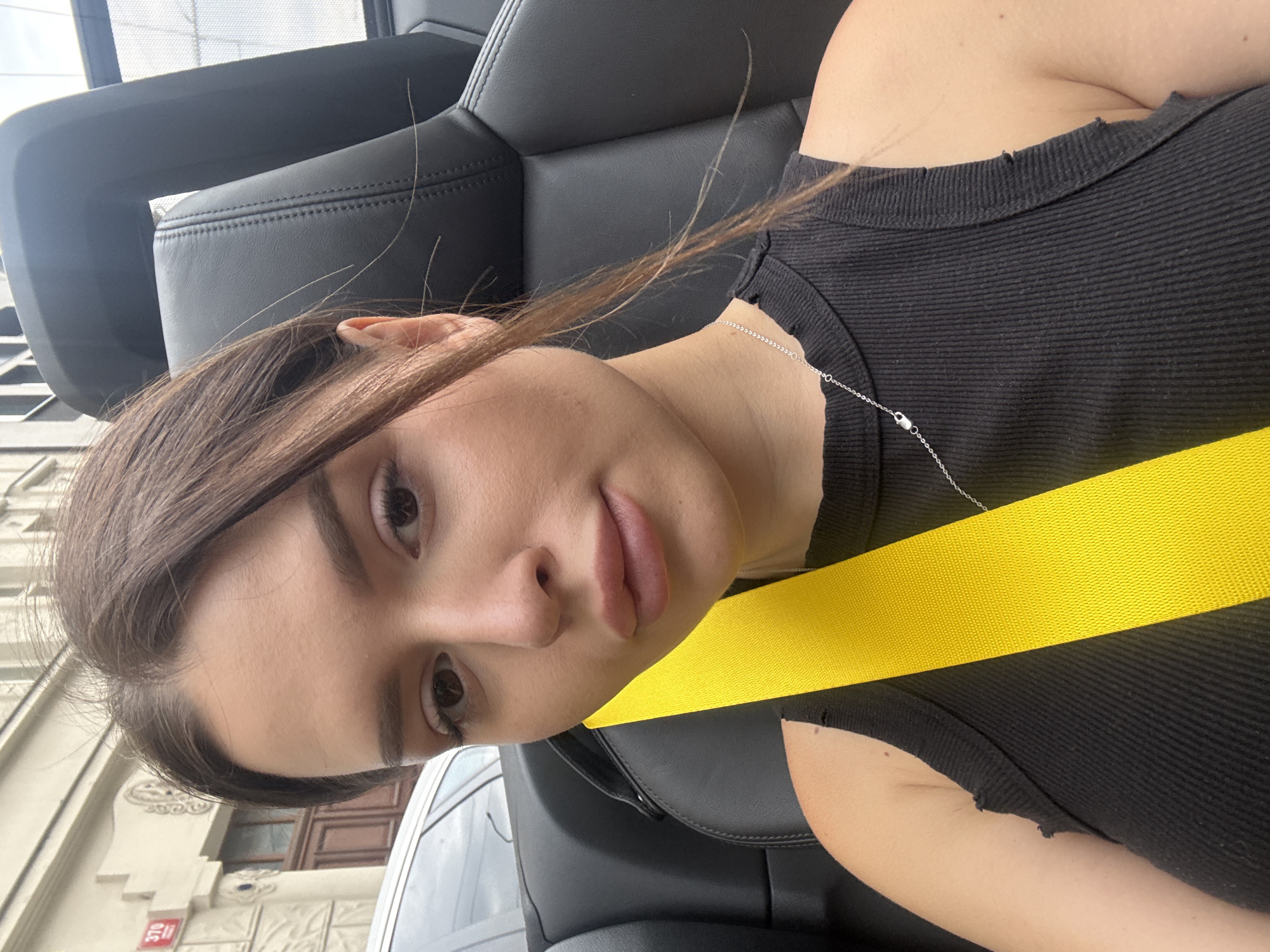 A young woman with brown hair pulled back is taking a selfie in the passenger seat of a convertible. She is wearing a black ribbed tank top and a silver necklace. A bright yellow seatbelt is across her chest. The interior of the car is black leather. In the background, a building with a red sign displaying "370" and a white and brown facade can be seen, indicating the location is Prague, Czechia.
