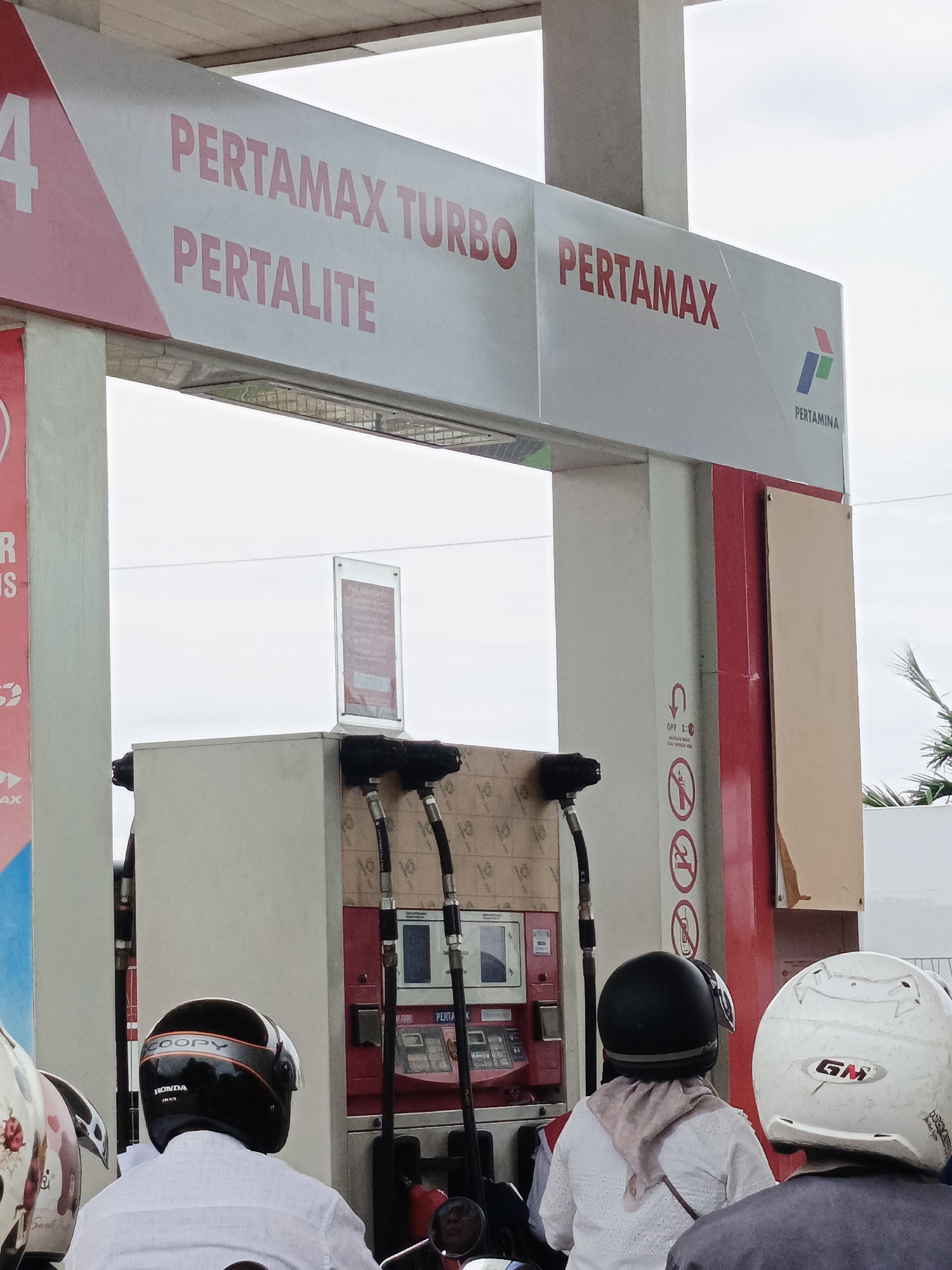 The image shows a Pertamina gas station in Cotbadatunong, Indonesia, with several motorcycles lined up to refuel. The gas station sign prominently displays "PERTAMAX TURBO," "PERTALITE," and "PERTAMAX" in red lettering against a white background, along with the Pertamina logo. Two individuals wearing helmets are seated on motorcycles, with the person in the foreground wearing a white shirt and a black helmet labeled "SCOOPY." Another person in a white helmet with "GM" branding is visible to the right. The gas pumps have hoses and nozzles ready for use. A red sign with cautionary symbols and text is mounted on a pillar, and a smaller information board is displayed near the pumps. The sky is overcast.