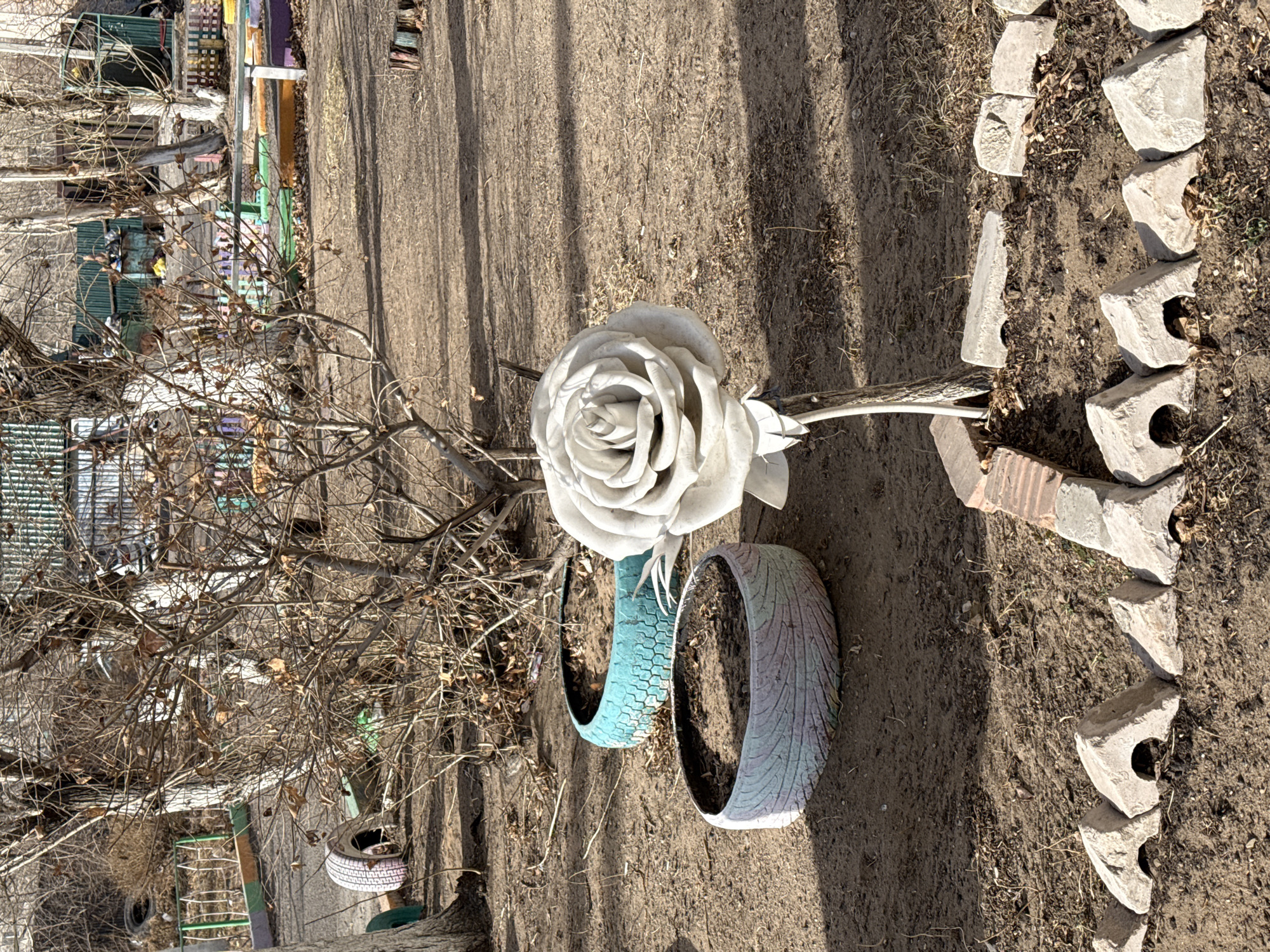 The image shows a ground-level view of an outdoor space, possibly in the city of Borzya, Russia. The scene includes a large, white rose sculpture on a stem, positioned among two painted tires used as planters, one blue and one grey. The ground is bare, and lined with grey concrete blocks and bricks. A tree trunk and branches are visible, with a structure in the background featuring green, white, and other colored elements. Additional tires are seen in the distance. The overall impression is one of a simple, rustic outdoor setting with hand-made decorative elements.