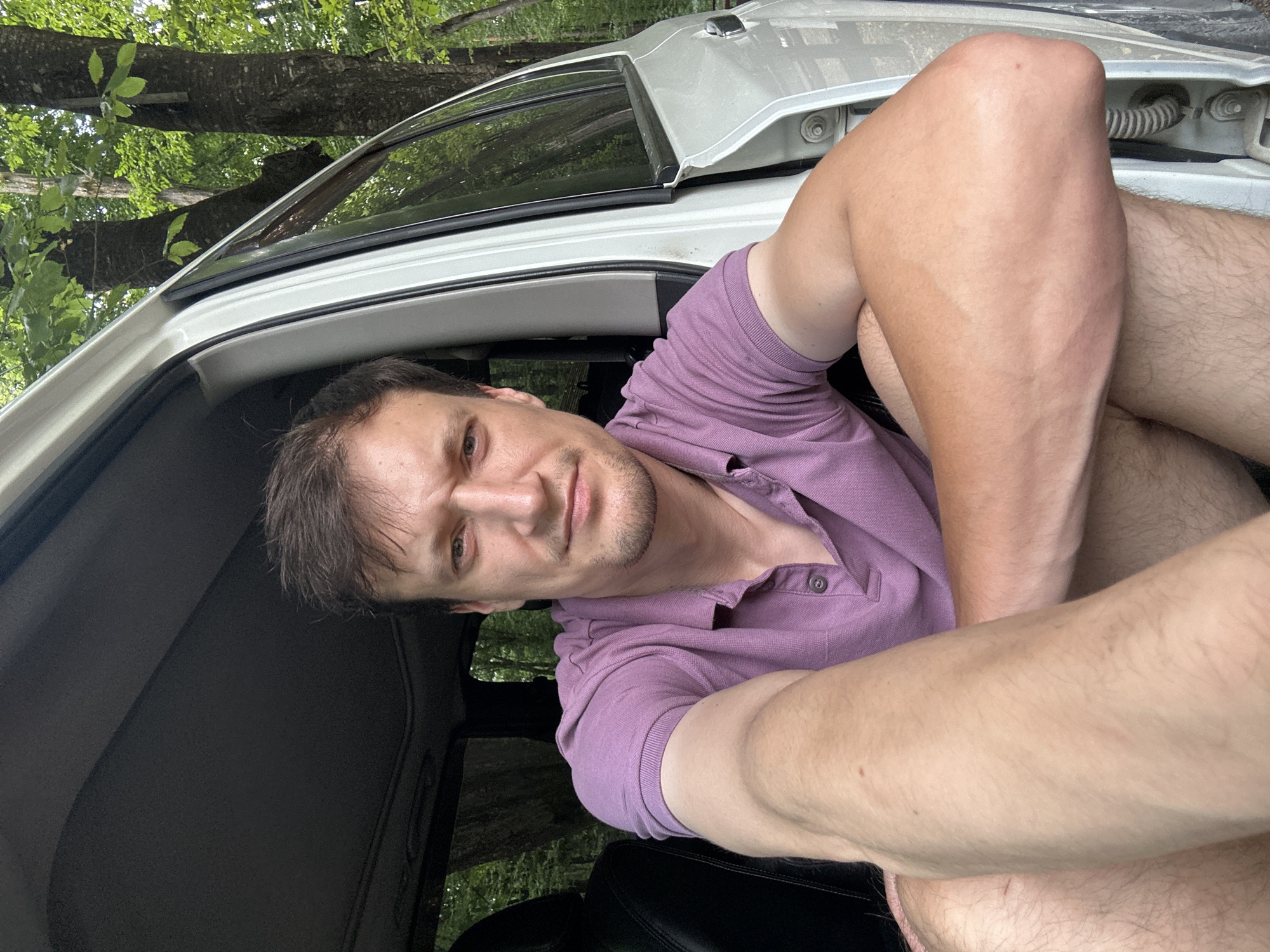 In Shatava, Ukraine, a man sits inside a vehicle with the doors open, in front of a wooded area. He is looking towards the camera, his left arm resting on his leg and his right arm bent. The man is wearing a purple polo shirt. The vehicle is white, and the interior is visible, as is the surrounding environment of trees and foliage.