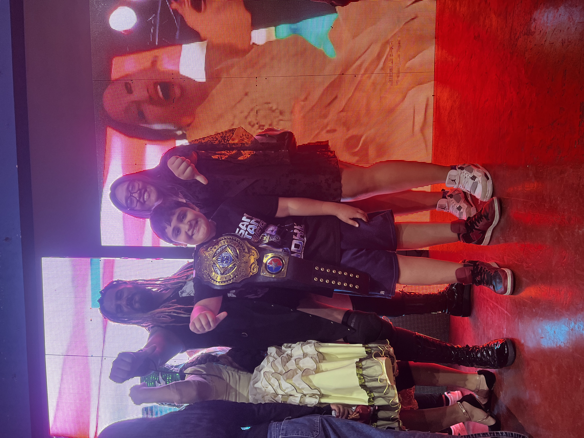 Arica, Chile: An indoor photo shows five individuals and part of a screen. A woman with glasses and dark hair is in the top right. Next to her is a boy wearing a championship belt. Below the boy is a man with dreadlocks. Further down are two additional people. The background consists of a screen displaying images and lights. The floor is red. People are interacting in the scene.