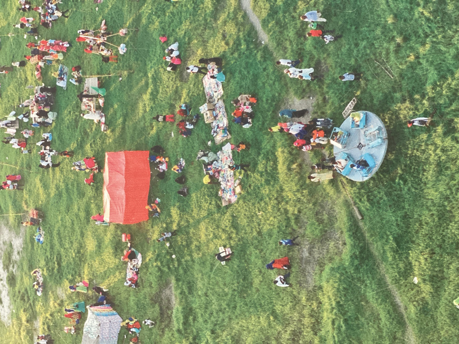 The image displays an aerial perspective of an extensive outdoor gathering on a verdant, uneven grassy field in Bububu, Tanzania. Over 100 individuals, consisting of adults and children, are distributed throughout the scene.
A light-colored, patterned tent is positioned in the upper-left section. Prominently in the central-left area is a large, rectangular structure covered in red material, likely a tent or stall. Multiple smaller groups of people are seated or standing around these structures.
The central and mid-ground portions of the field host numerous informal market displays. Goods, including assorted clothing, boxed items, and various small products, are laid out on ground coverings such as blankets and tarps. Individuals are observed engaging in commercial activities, including browsing, selling, and purchasing. Other groups are seated on the grass, interacting or observing. One person in the mid-left field is holding a stringed object, possibly a kite.
In the lower-central foreground, a light-blue carousel ride with multiple occupied individual seats is operational. Surrounding this attraction are additional clusters of adults and children. A distinct dirt path winds through the lower right quadrant of the field. The scene is illuminated by natural light, suggesting daytime.