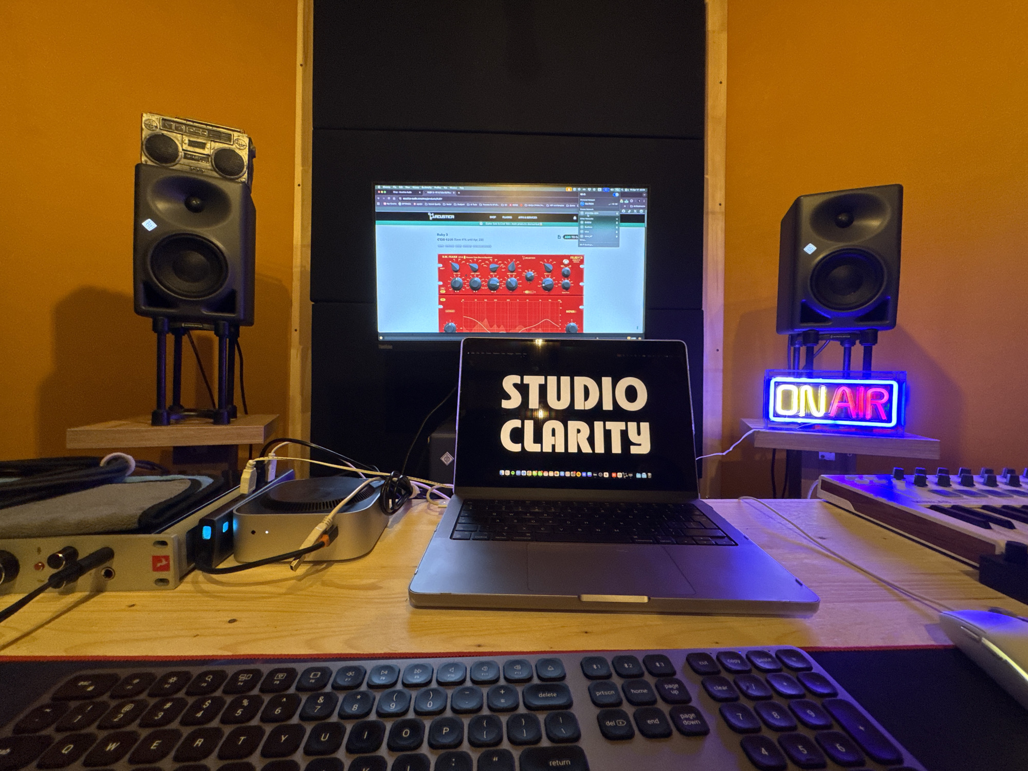 The image shows a recording studio setup. A laptop with the words "STUDIO CLARITY" on the screen is centrally placed. In front of the laptop, there's a keyboard and a mouse on a red mousepad. Behind the laptop, a monitor displays a screen with audio plugins visible. On either side of the monitor, there are studio monitor speakers on stands. The left speaker has a boombox placed on top of it. A neon "ON AIR" sign is placed beside the right speaker. Audio interface equipment is visible on the left side of the table. The background walls are painted in shades of orange and wooden panels. The image was not taken in the city of Plovdiv, Bulgaria, as there are no indicators of location in the image.