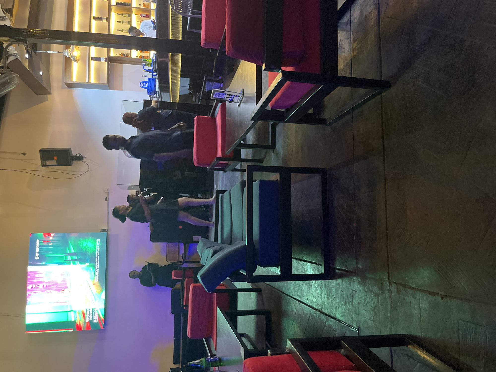 The image depicts an indoor scene at a bar or lounge, likely in Abuja, Nigeria, characterized by ambient lighting and modern furnishings. In the background, a large LED screen displays a colorful, abstract visual. Several individuals are visible. Two women in black dresses stand near a DJ booth or sound system, one of whom is holding a tablet. Further back, a man in a black shirt appears to be walking. To the right, a bar area is illuminated by warm lights, with bottles of liquor displayed on shelves. A bartender in a white shirt is attending to the bar. In the foreground, a seating area features contemporary furniture, including a long, dark-colored sofa with blue cushions and several armchairs with vibrant red cushions. Dark wooden tables with metallic frames are placed among the seating. The floor is made of dark wooden planks, with areas illuminated by green light, suggesting mood lighting.