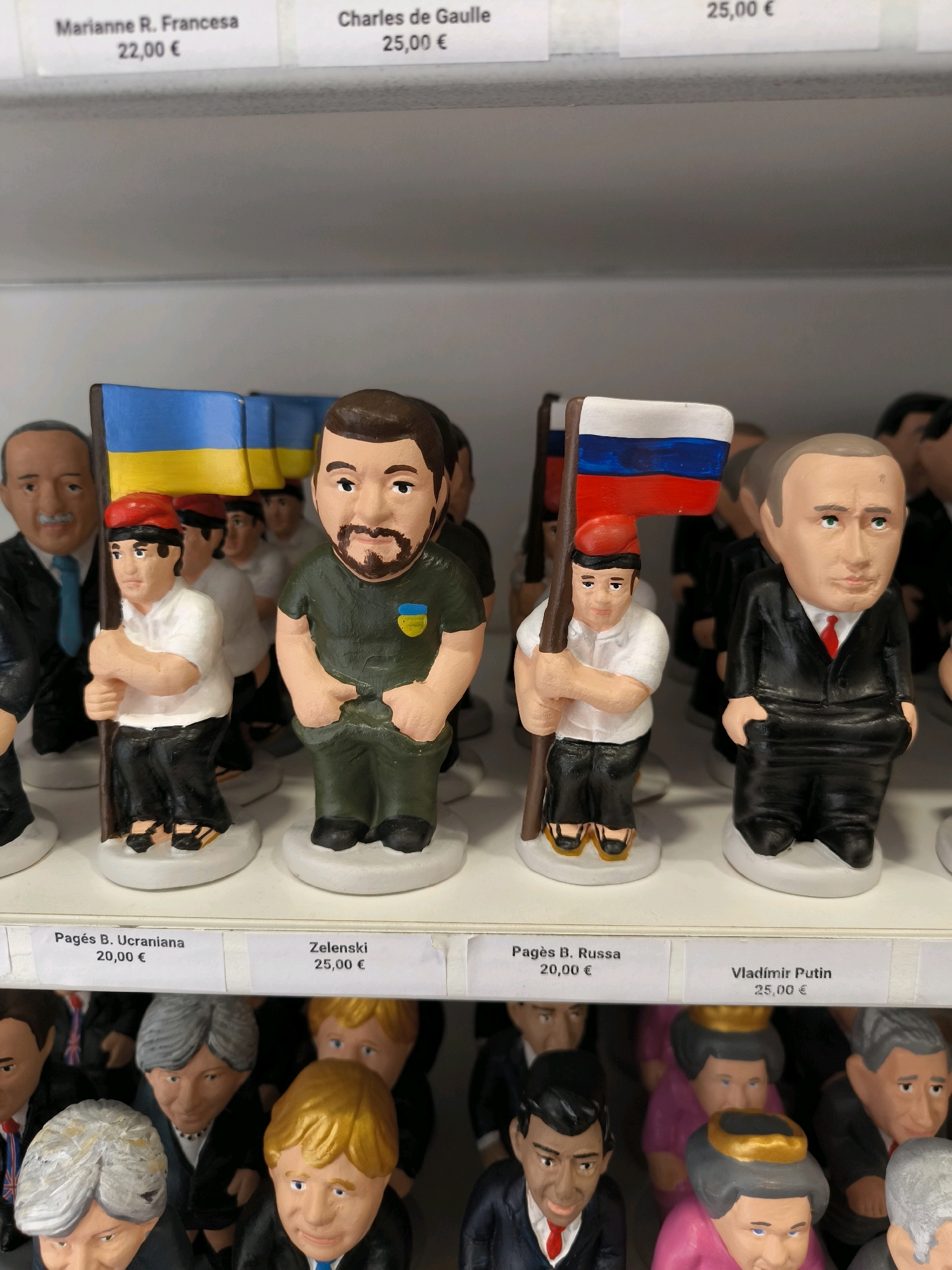 The image shows a shelf display of small, caricature-like figurines of political figures. In the foreground, there are two figures prominently displayed. The first figure on the left is wearing a green military-style uniform with a Ukrainian flag emblem on the chest and holding a Ukrainian flag. The second figure on the right is dressed in a black suit with a red tie and is holding a Russian flag. Both figures have exaggerated features and are standing on white circular bases. Behind these figures, other figurines of political leaders are visible.
Below the figurines, there are price labels. One label reads "Pagès B. Ucraniana 20,00 €" and another reads "Zelenski 25,00 €". Further to the right, a label reads "Pagès B. Russa 20,00 €" and "Vladimir Putin 25,00 €". In the top portion of the image, another row of labels is partially visible, one reading "Marianne R. Francesa 22,00 €" and another "Charles de Gaulle 25,00 €". The display is set against a plain, neutral background, likely a store shelf. The lighting is even and diffused, suggesting an indoor environment. The overall scene depicts merchandise for sale in a retail setting. This scene is located in the city of Bilbao, Spain.