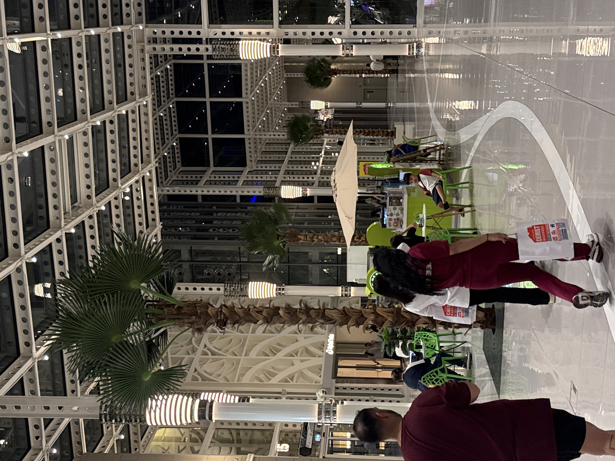 The image depicts the interior of the Dubai Mall in Dubai, United Arab Emirates. The scene captures a wide, well-lit atrium area with high ceilings supported by a metallic grid structure. Large palm trees are strategically placed throughout the space, adding an element of nature to the modern architecture. Several people are visible in the foreground and midground, some walking and others seated at tables.
In the foreground, a man wearing a burgundy t-shirt and black shorts is walking away from the camera. To his right, a woman in a maroon outfit walks towards the right, carrying a white shopping bag. Behind her, another woman in a white top and black pants walks in the same direction.
In the midground, several small cafe-like seating areas with bright green chairs and small tables are set up. One area features an umbrella providing shade over a table, with what appears to be a small food or drink kiosk behind it. A young boy is seated at one of these tables, looking towards the left. Other individuals are seen sitting or standing in these seating areas, interacting or engaged in individual activities.
The flooring is a highly reflective polished surface, mirroring the lights and the architectural elements of the mall. The overall ambiance suggests a bustling, contemporary shopping and leisure environment.