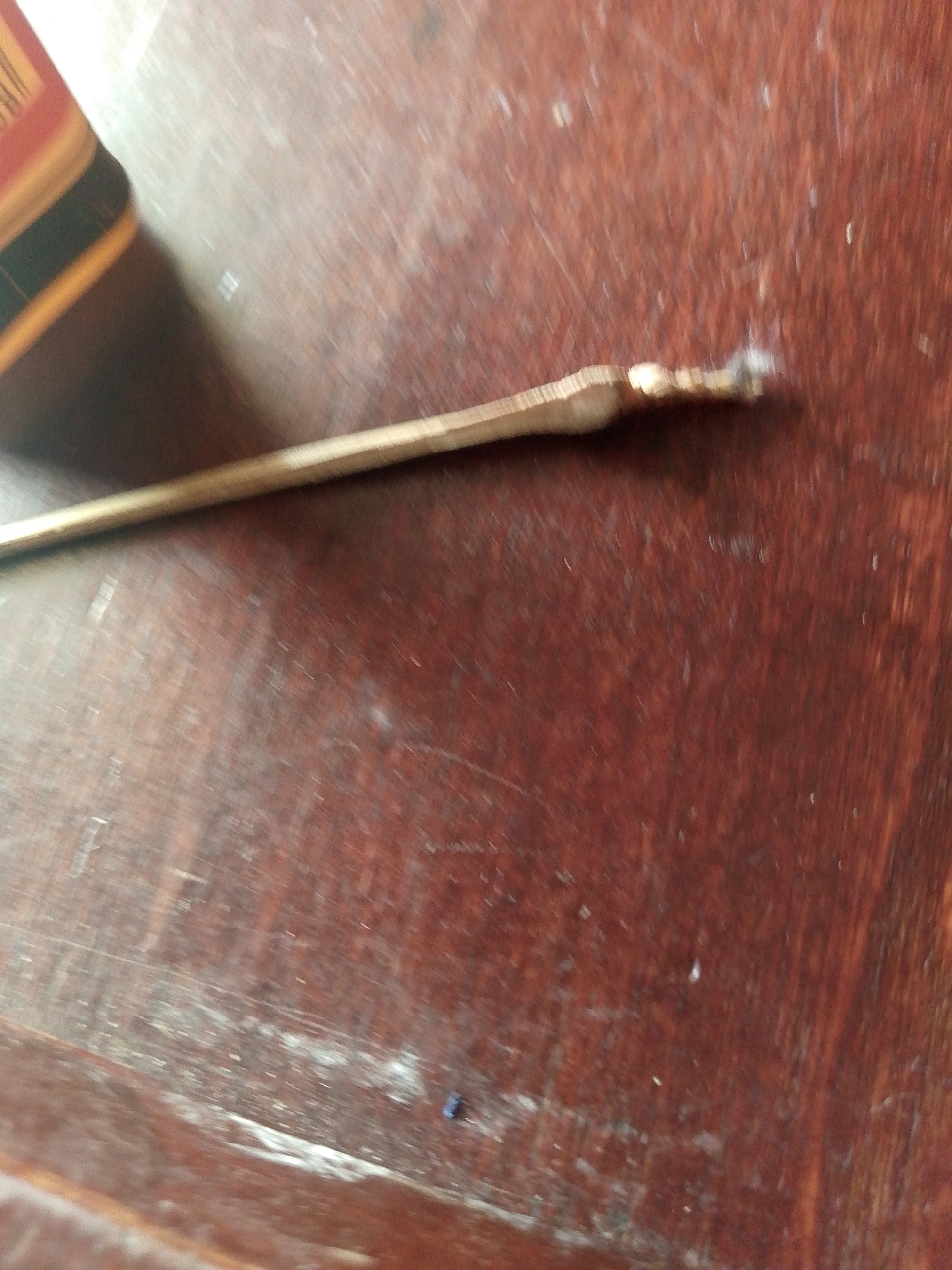 A close-up, eye-level shot shows a brass poker lying on a reddish-brown wooden surface. The poker is positioned diagonally from the bottom left to the top right of the frame. The surface has a visible wood grain pattern and appears to be dusty or have some light-colored debris scattered across it. In the background, on the left side of the image, a portion of a book or similar object with colored bands of yellow, red, and black is visible. The lighting suggests an indoor setting, possibly on a table. The image does not contain any individuals or provide any specific location details within Jauro Yinu, Nigeria.