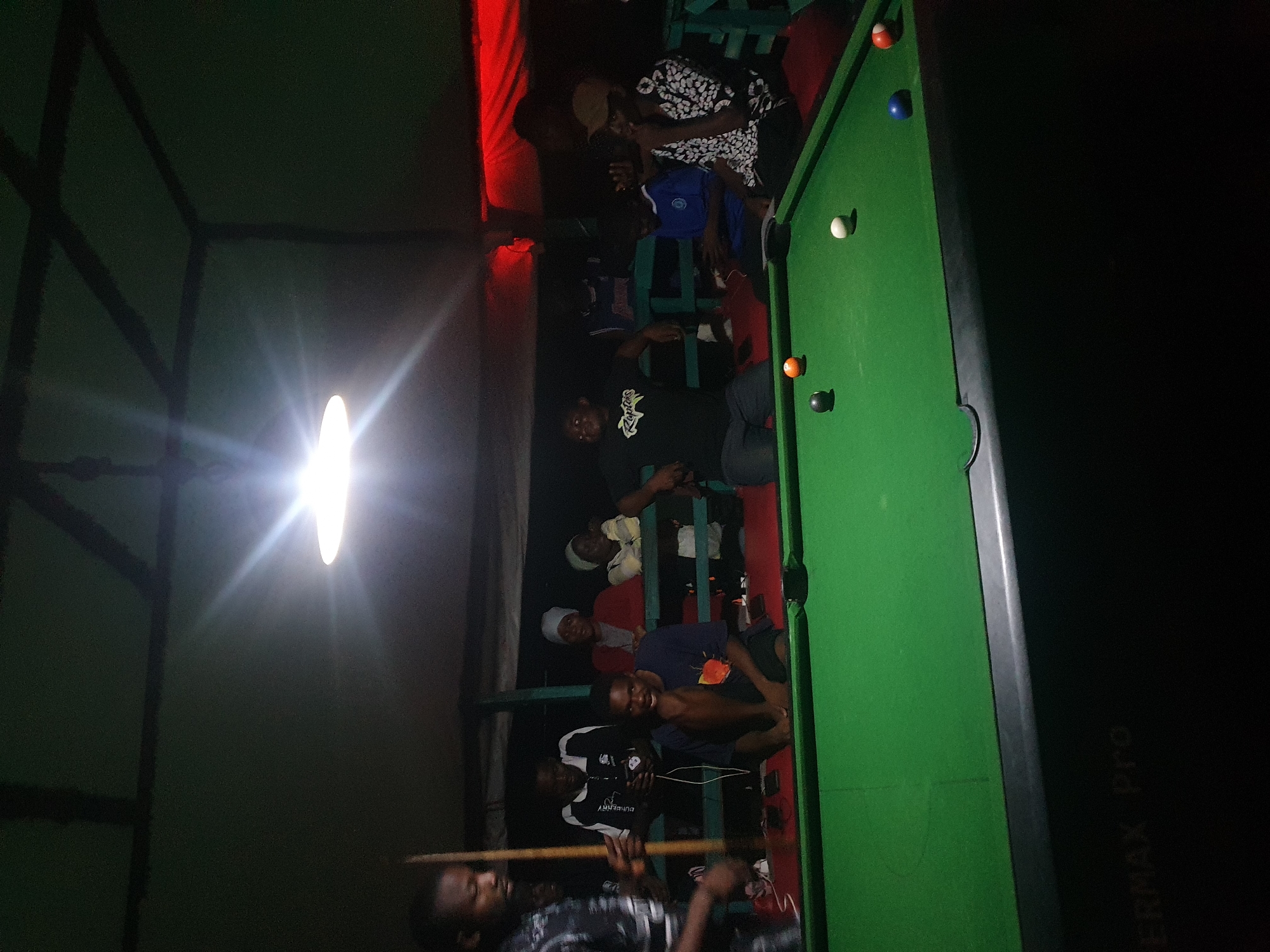 A group of young men are gathered around a pool table in a dimly lit indoor space. The scene appears to be a casual gathering, possibly a pub or a recreational center, given the presence of the pool table and the relaxed postures of the individuals. A bright light source, likely a bare bulb, illuminates the left side of the image, casting long shadows and creating a stark contrast with the darker areas.
The individuals are dressed in casual clothing, with some wearing t-shirts and others in more traditional attire. They are positioned around the pool table, with some actively engaged in playing or observing the game. One person is holding a pool cue, suggesting an ongoing game. The pool table itself is green with several balls visible, including a white cue ball, an orange ball, and a black ball. The red edge of the table is also visible.
The setting is somewhat rustic, with exposed beams or structural elements visible in the background on the left. The overall atmosphere is informal and social. The time of day cannot be definitively determined, but the artificial lighting suggests it could be evening or night. There are no obvious cues about the weather.
Visible text includes "BURBERRY" on one of the individuals' shirts and "PERMAX Pro" on a dark object on the right side of the image, possibly a speaker or electronic device. The emotions conveyed are a mix of concentration and camaraderie among the people. The location context of Lokoja, Nigeria, points to a specific cultural and geographical setting for this gathering.