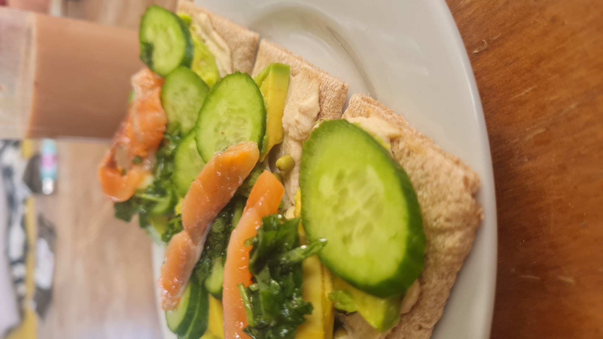 The image captures a close-up, high-angle view of a freshly prepared meal, presented on a white ceramic plate. The food appears to be an open-faced sandwich or toast made with whole wheat or flatbread, likely cut into rectangular portions.
The bread pieces are generously spread with a light-colored, creamy substance that strongly resembles hummus. Abundantly layered on top are various fresh ingredients: multiple slices of vibrant green cucumber, creamy green avocado slices, and several strips of orange-colored fish, consistent with smoked salmon. Interspersed among these toppings are small clusters of dark green leafy herbs, possibly parsley or dill, and a few tiny, round green elements that could be capers or peas. The ingredients are neatly arranged, indicating careful preparation.
The plate rests on a warm, light brown wooden surface, likely a table. In the blurred background to the left, a tall, brown-colored container or bottle is faintly visible, suggesting a drink accompanying the meal. The lighting is bright, highlighting the freshness and textures of the food.
The scene depicts a meal ready for consumption, likely breakfast or lunch. There are no people visible, nor any discernible text or branding. The fresh ingredients and the presence of hummus align with culinary traditions found in the Mediterranean region, fitting the given location context of Lachish Regional Council, Israel. The setting appears to be indoors, possibly a home or a cafe, during daylight hours.