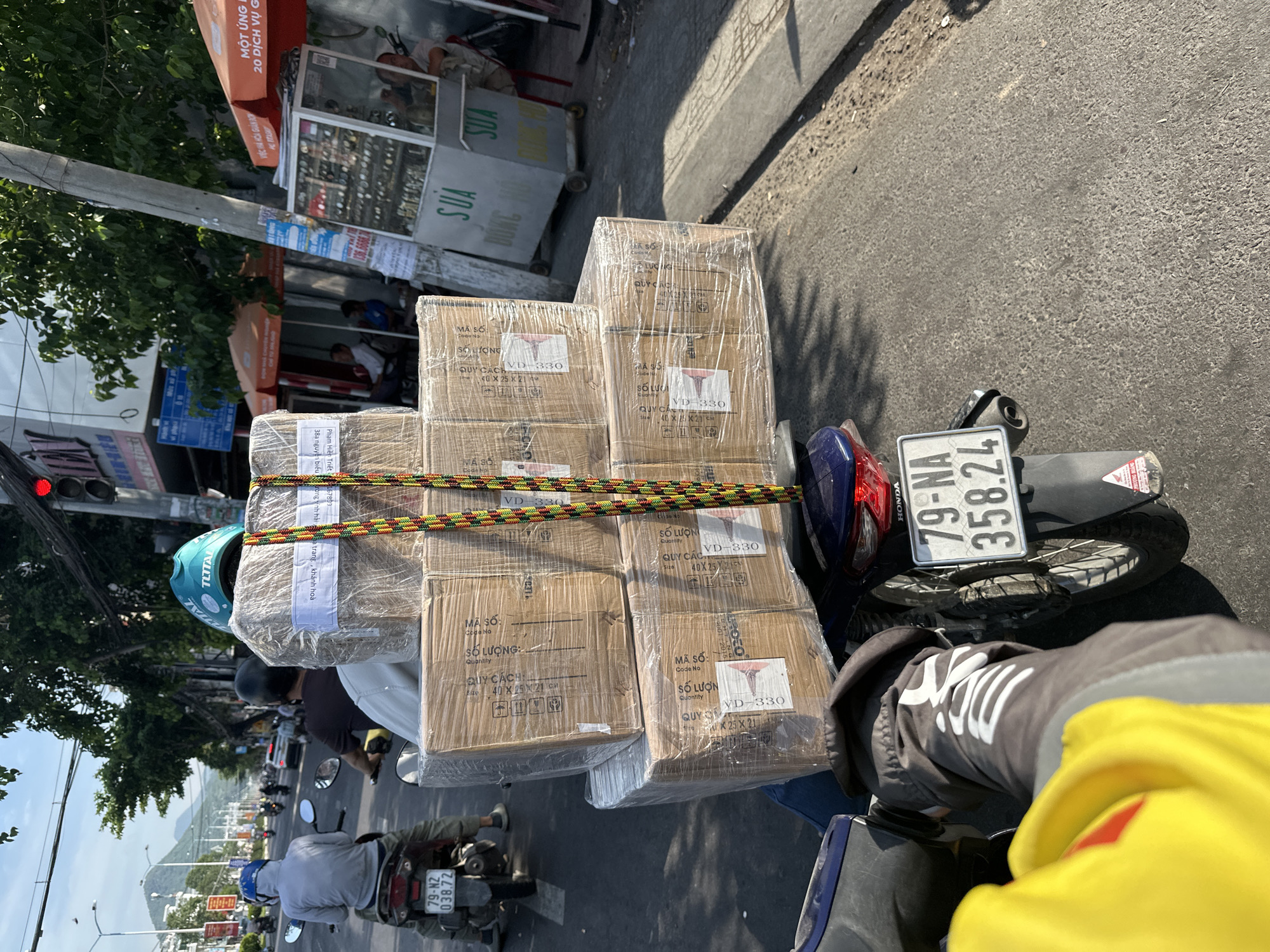 In a street scene in Bac Nha Trang Ward, Vietnam, a person riding a motorcycle is captured from a rear-facing perspective. The motorcycle is loaded with several large cardboard boxes, tightly wrapped in clear plastic and secured with colorful ropes. The license plate of the motorcycle reads "79-NA 358.24". The rider is wearing a dark jacket with white writing on the sleeve. In the foreground, a portion of a bright yellow object, possibly a container, is visible. The background shows a street with other motorcycles and vehicles, as well as buildings and trees, indicating an urban environment. A traffic light is visible at the top of the frame. The sunlight suggests it is daytime.