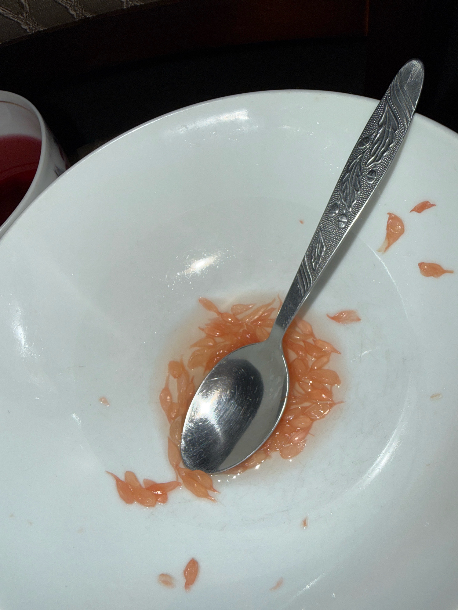 A silver spoon rests in a white bowl, surrounded by remnants of grapefruit segments. The bowl is on a dark brown surface, possibly a table. To the left of the bowl, a portion of a white cup with a red liquid is visible. The scene appears to be a domestic setting. There is no information about the location in the city of Zavolzhye, Russia, or any people present.