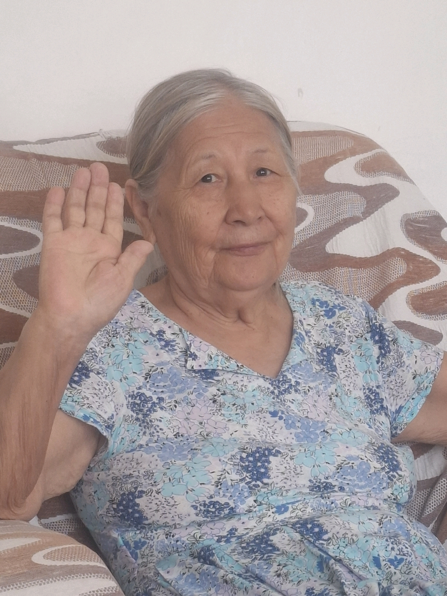 An elderly East Asian woman with graying hair is seated in a chair, her left arm raised with her palm facing forward, as if waving or signaling. She is wearing a blue and white floral patterned blouse. The chair appears to be upholstered with a patterned fabric. The background is a plain, light-colored wall. The scene is indoors. There is no discernible indication of the specific location within the city of Almaty, Kazakhstan.