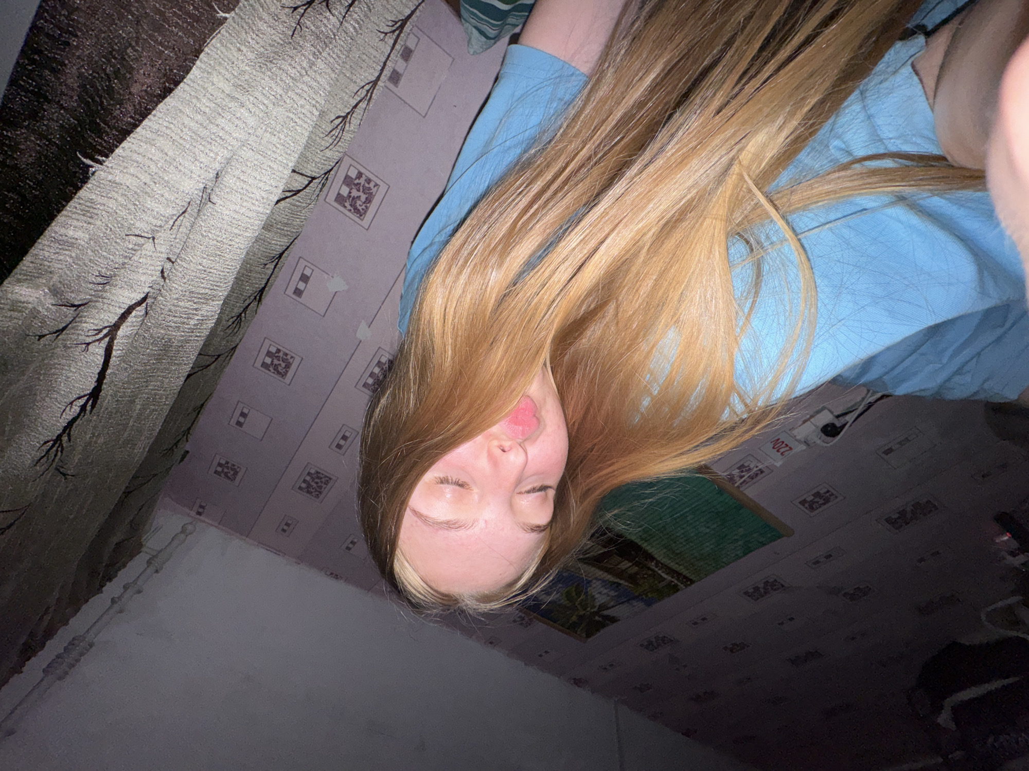 The image depicts a young woman with long, blonde hair wearing a light blue shirt. She is lying down, possibly on a bed, with her face turned towards the camera. Her eyes are closed and her lips are pursed. The background includes a wall with a patterned wallpaper and a grey curtain. The environment appears to be a room in Perm, Russia, indicated by the 220V indicator (voltage for Russia) on the ceiling and a photo of a palm tree. The overall lighting is dim.