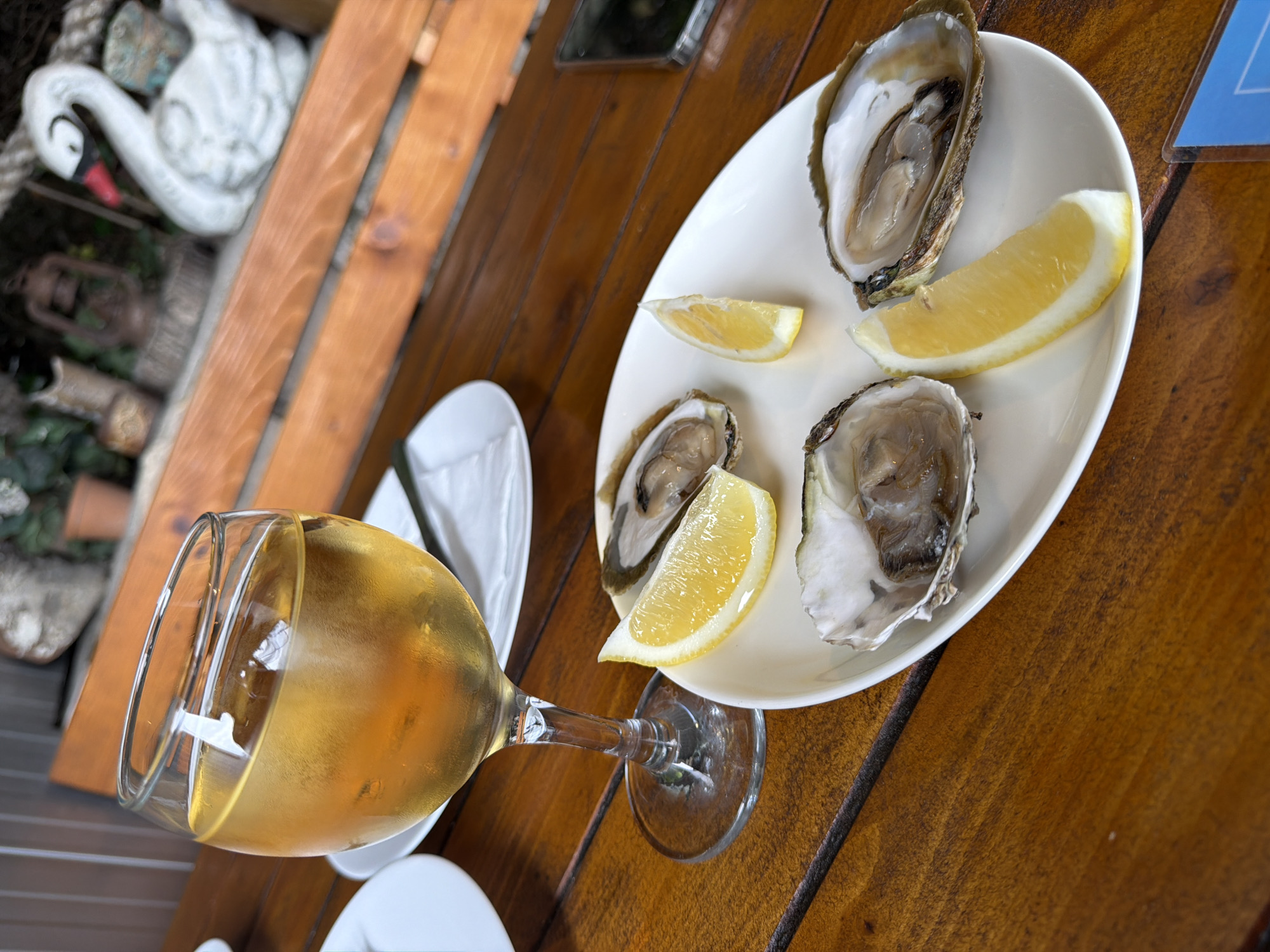 On a wood table at an unidentified location, there is a wine glass full of amber-colored liquid and a white plate with three open oysters and lemon wedges. A white swan statue can be seen in the background, along with additional plates and a cell phone.