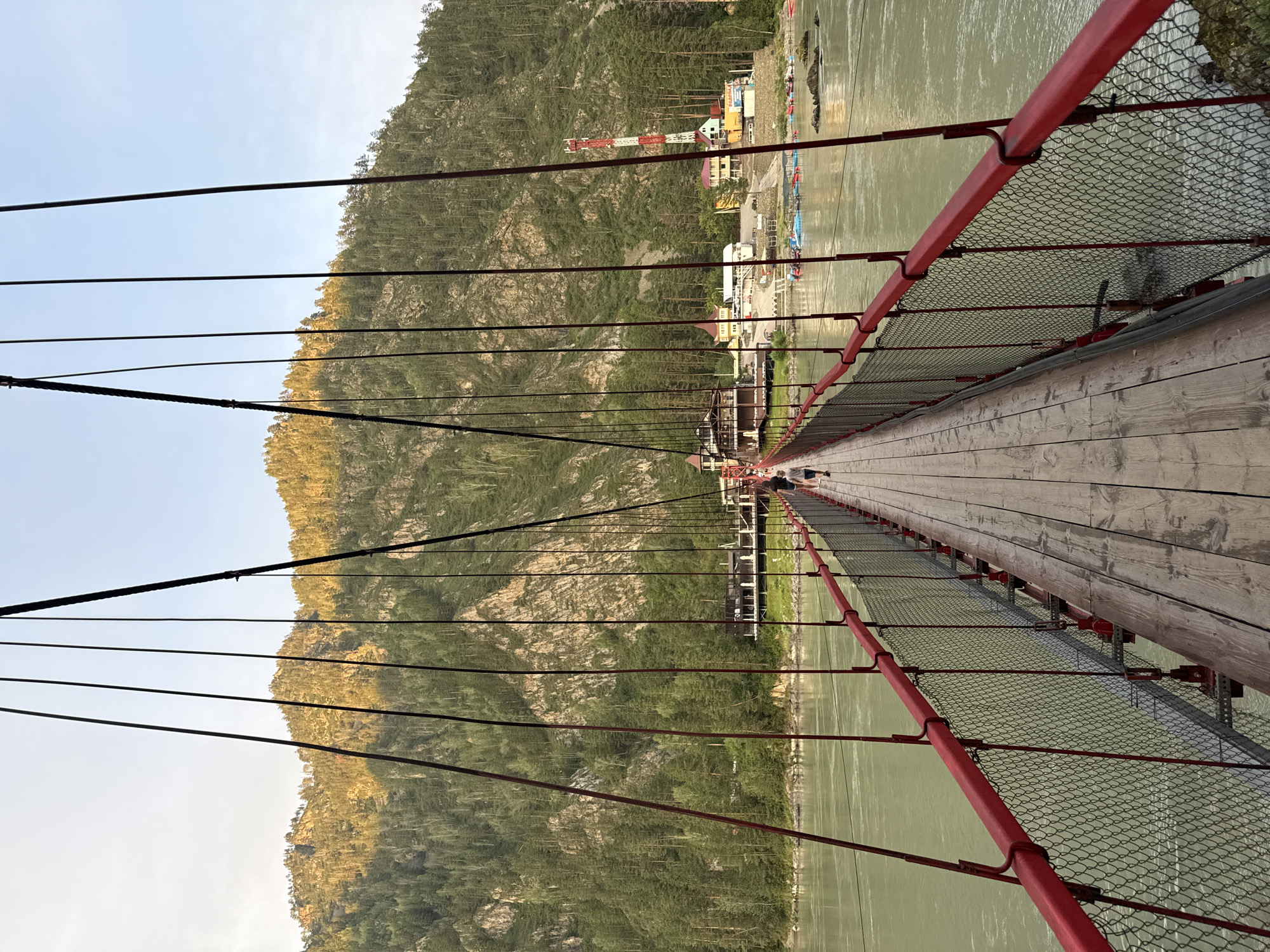 The image captures a view from a suspension bridge in the village of Barangol, Russia, spanning a wide, greenish-gray river. The bridge has a wooden plank walkway and red metal railings with chain-link fencing. Several tall, thin cables, some black and some red, extend upwards from the bridge, supporting its structure. Two individuals, one appearing to be a child and the other an adult, are walking away from the camera on the bridge.
In the background, across the river, is a steep, forested mountainside bathed in the warm light of the setting or rising sun, highlighting the green of the trees and the rocky outcrops. Along the riverbank, a settlement is visible, featuring several buildings with peaked roofs, a red and white communications tower, and numerous small boats, some appearing to be rafts or kayaks, docked near the shore. The sky is clear and blue, with a hint of haziness near the horizon.