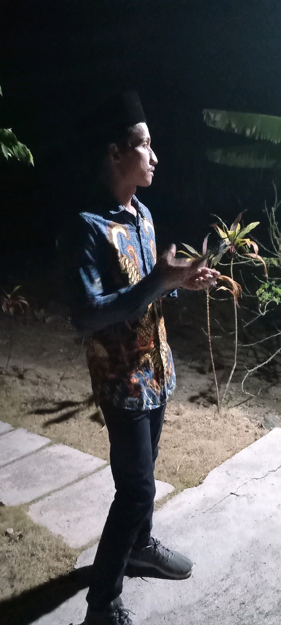In Kedungmulyo, Indonesia, a young man stands outdoors at night. He is wearing a black peci hat, a blue and orange batik shirt, and black pants. His right hand is raised and slightly cupped, as if holding something or gesturing. The background is dark, with some foliage visible, including a plant with reddish-brown leaves to his right and a hint of green leaves to his left. The ground appears to be a mix of dirt and paved sections, with concrete slabs forming a pathway. The lighting suggests artificial illumination, casting shadows on the ground.