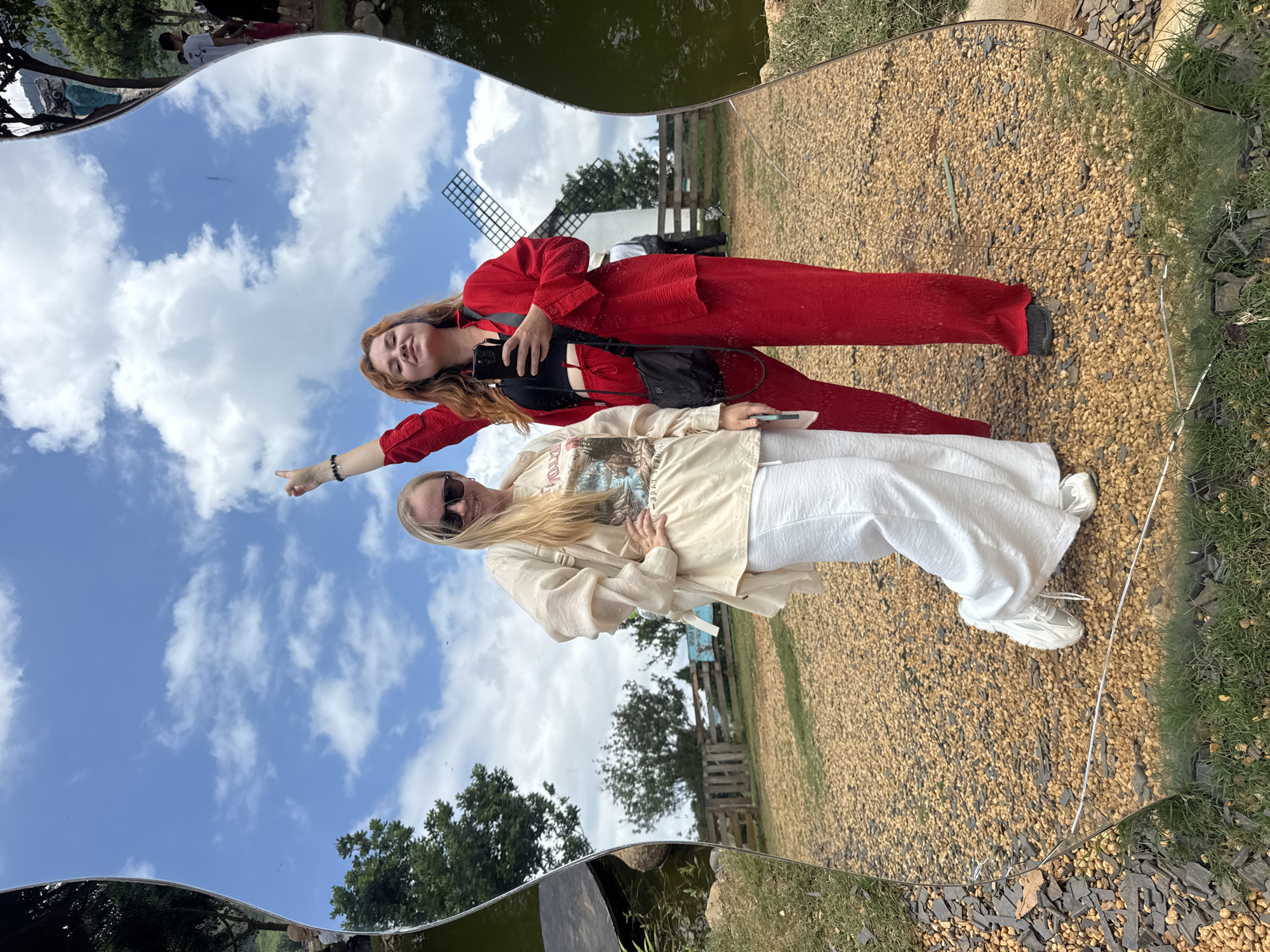 An outdoor, eye-level, full-shot photograph captures the reflection of two women posing in a large, irregularly shaped, possibly wavy mirror situated in Thôn 5, Vietnam. The mirror, which shows minor cracks and a horizontal scratch near its bottom edge, distorts the reflected images, particularly along its curved borders.
On the left side of the reflection is a woman with blonde hair, wearing sunglasses, a light-colored long-sleeved top or jacket over a graphic t-shirt, white wide-leg trousers, and white sneakers. Her right hand rests on her hip, and she smiles towards the viewer.
To her right stands a woman with reddish-brown hair, smiling and looking towards the camera. She wears a red textured long-sleeved collared shirt open over a black crop top, matching red textured wide-leg trousers, and dark flat shoes. She carries a black crossbody bag. Her left arm is raised with the index finger pointing upwards, while her right hand holds a black smartphone.
The background, visible both reflected in the mirror and around its edges, depicts a bright blue sky with scattered white cumulus clouds. A prominent white windmill with dark blades is reflected behind the woman in red. The ground is largely covered with light-brown pebbles or gravel, with patches of green grass visible along the bottom edge where the mirror meets the terrain. A wooden fence runs horizontally across the mid-ground, behind which are green trees and a partially visible blue sign. On the far left edge of the mirror's reflection, a dark body of water with surrounding foliage is visible. Faint reflections of other individuals are discernible on the extreme curved edges of the mirror, suggesting a public outdoor setting.