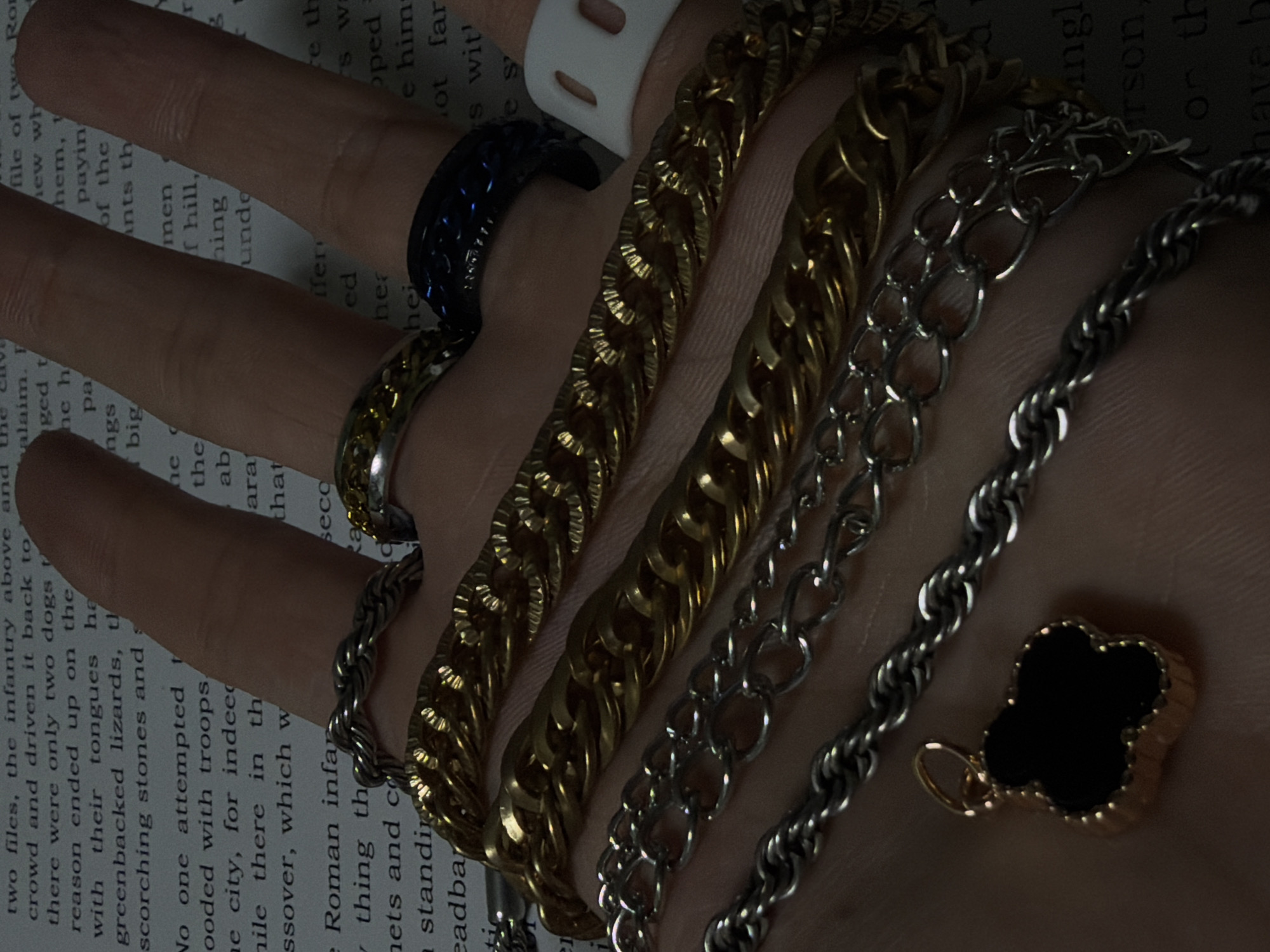 In an indoor environment with poor lighting, a hand adorned with multiple jewelry items rests on a book. The hand displays four rings: a braided silver ring on the index finger, a gold and silver ring on the middle finger, a dark blue ring on the ring finger, and a white plastic ring on the pinky finger. Several chains are layered on the hand and wrist, including a thick gold chain, a thinner gold chain, a silver chain, and a rope-style silver chain. A clover-shaped pendant with a black center and gold trim is attached to one of the chains. The book's pages are visible in the background, with text suggesting a historical or literary context. The overall scene is dark and muted, with the jewelry being the most prominent and well-lit elements.