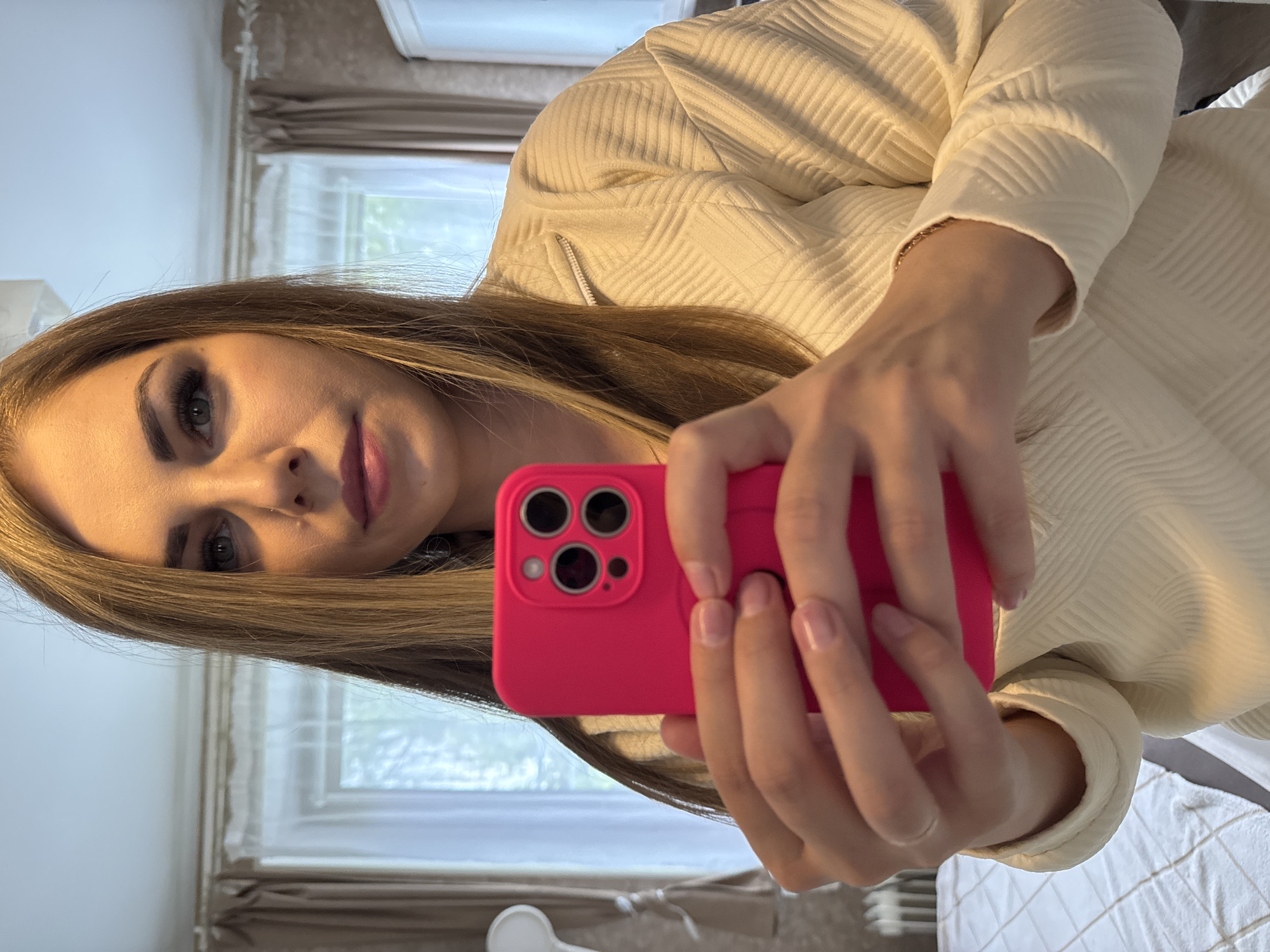 A woman is taking a selfie with a pink smartphone. She has light brown hair and is wearing a cream-colored textured jacket. A window with curtains is visible in the background. The location is not discernible from the image.