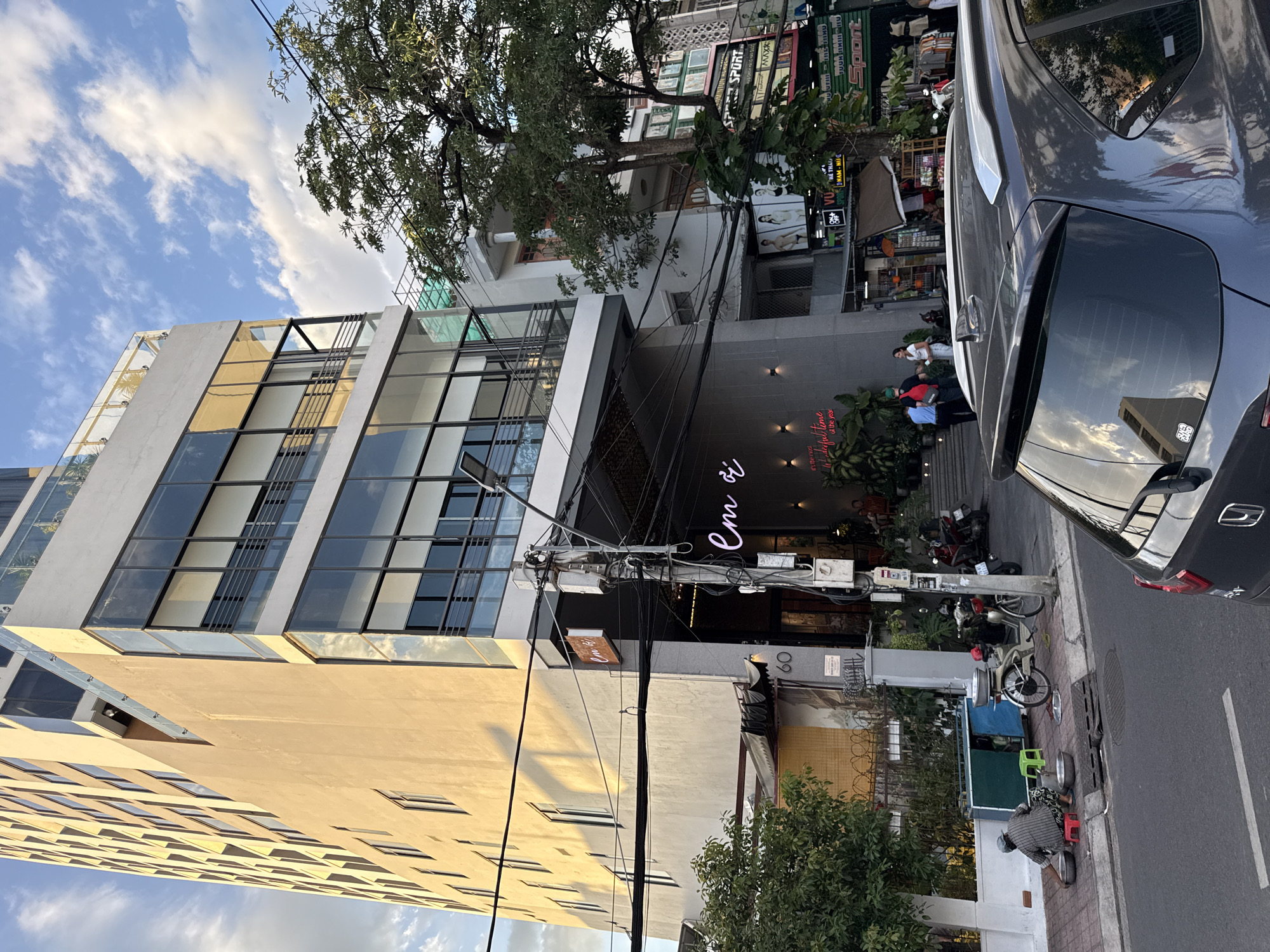 The image shows a street scene in Nha Trang, Vietnam. A tall, modern building with many windows dominates the left side of the frame. The building appears to be a hotel or apartment complex, with a lower section featuring a business with a neon sign that reads "Em ơi."
In the foreground, a dark-colored SUV is parked, partially obscuring the street. On the sidewalk to the left, a person is sitting on a red stool next to a blue and green cart, washing bowls in large metal basins. Several motorcycles are parked nearby.
Further down the street, other businesses with signage are visible, including what appears to be a sports apparel store with the word "SPORT" displayed prominently. A large tree with dense foliage partially obscures some of the buildings on the right. The sky is blue with scattered clouds, and the sunlight casts shadows on the buildings and street. The overall scene depicts a typical urban environment with a mix of residential and commercial activity.