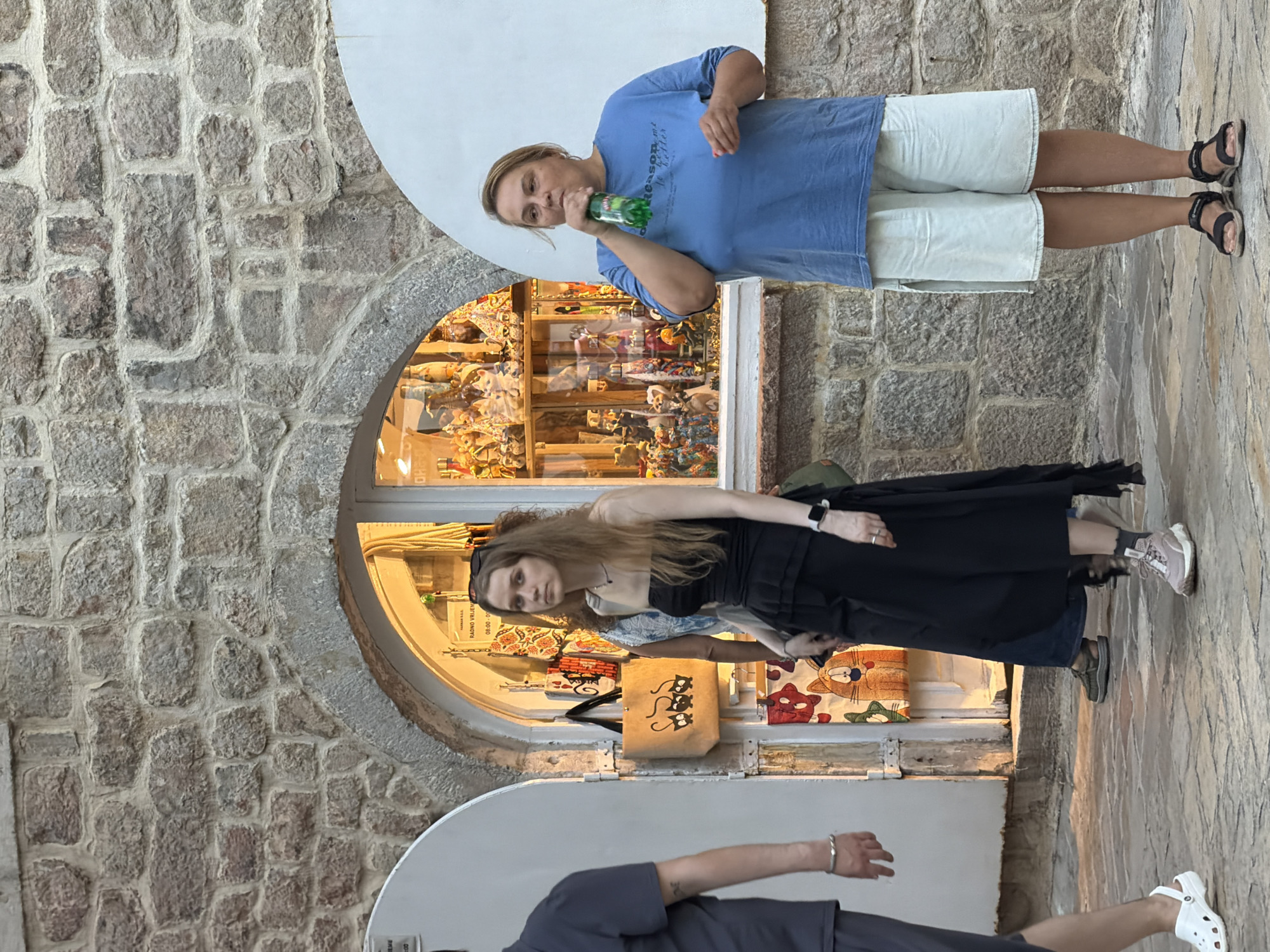 In Budva, Montenegro, a woman in a blue t-shirt and light-colored shorts drinks from a green bottle, standing on a stone-paved street. To her left, a young woman in a black strapless dress walks past a shop window displaying various figurines. Behind her, another person is partially visible. To the far left, the legs of a person in a grey shirt and shorts are seen walking away from the camera. The shop's exterior features a stone wall with an arched opening leading to the window display. The scene is illuminated by warm, artificial light emanating from the shop.