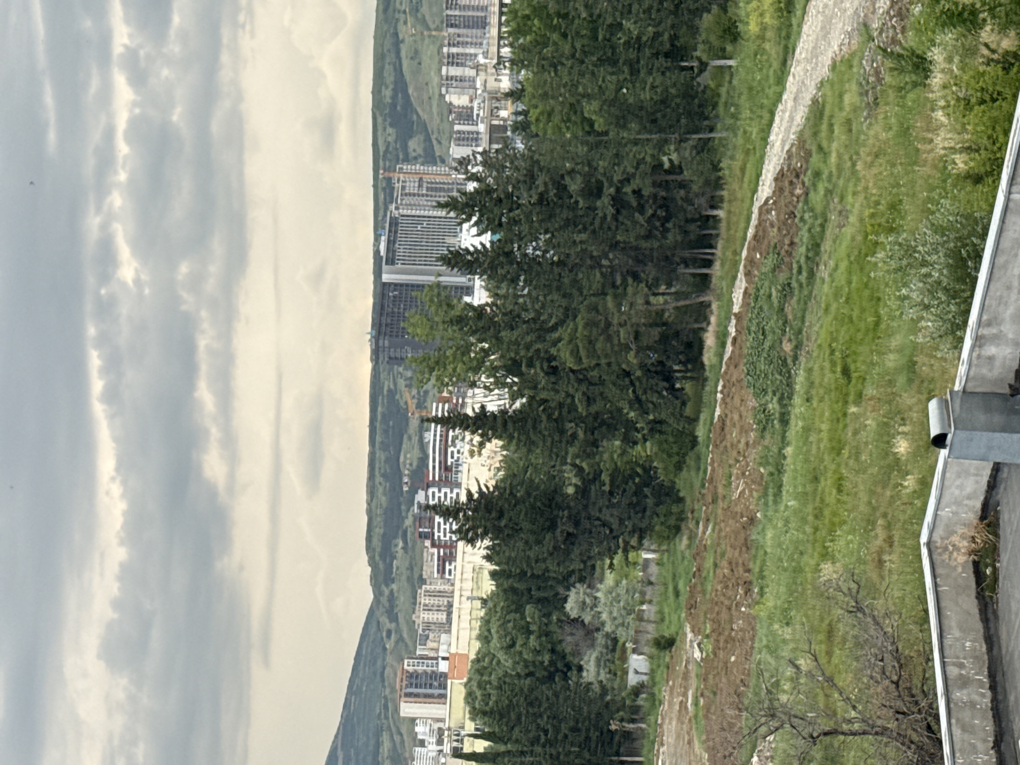 This media file shows a view of a city from a raised vantage point, likely a hill or elevated structure. The scene is dominated by a panorama of buildings, interspersed with lush green trees and rolling hills in the background. The sky is overcast with heavy clouds, suggesting a cloudy or possibly rainy day. The image is oriented vertically, with the cityscape stretching out horizontally across the frame.
In the foreground, a textured wall made of stones and dirt slopes downwards, bordered by green grass and some sparse vegetation. Part of a concrete structure with a metal downspout is visible at the very right edge of the image, indicating the photographer's elevated position.
The cityscape features a mix of modern and possibly older architecture, with numerous buildings of varying heights and styles. Some buildings appear to be under construction, with visible scaffolding. The dense urban sprawl suggests a bustling city environment. The overall mood conveyed by the image is somewhat somber due to the cloudy weather, but also hints at the vastness and complexity of the urban landscape.
The location is identified as Tbilisi, Georgia. While there are no people or specific activities depicted, the scene captures a typical urban vista under a cloudy sky. No visible text is present in the image.