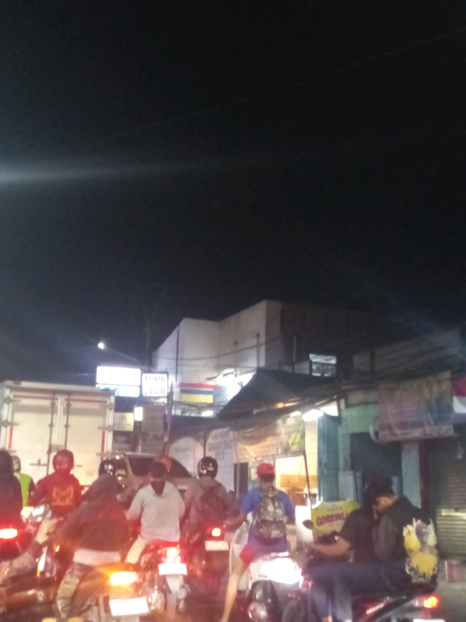 The image depicts a nighttime scene of heavy traffic congestion on a city street in Bekasi, Indonesia. Several motorcycles and a truck are visible. Most individuals are wearing helmets, suggesting a typical urban environment where motorcycle use is prevalent. The presence of streetlights and illuminated signs indicates a commercial or residential area. The dark sky suggests it is late evening or night. One sign prominently displays the words "GORENGAN" which translates to fried snacks, indicating a food stall or vendor is present. The overall atmosphere is one of a busy, bustling city at night, with traffic being a significant element of the scene.
