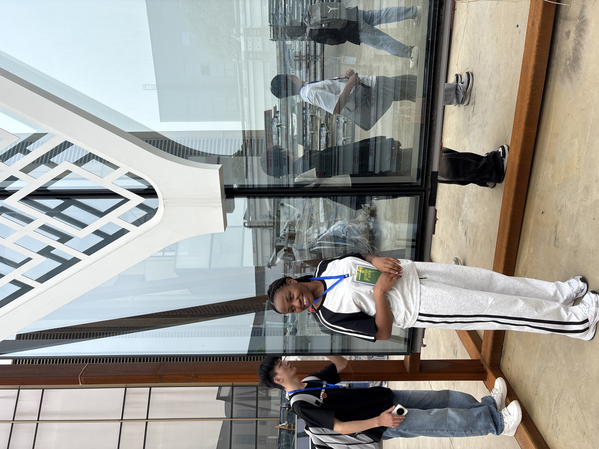 Two young people pose for a photo in front of a modern architectural structure, possibly a building, located in Chengdu, China. The structure features a geometric design and large glass panels, reflecting multiple individuals and internal details of the building. The woman is holding a book in her hands. Both individuals are wearing lanyards around their necks. The environment includes architectural elements with a mix of modern and possibly weathered materials.