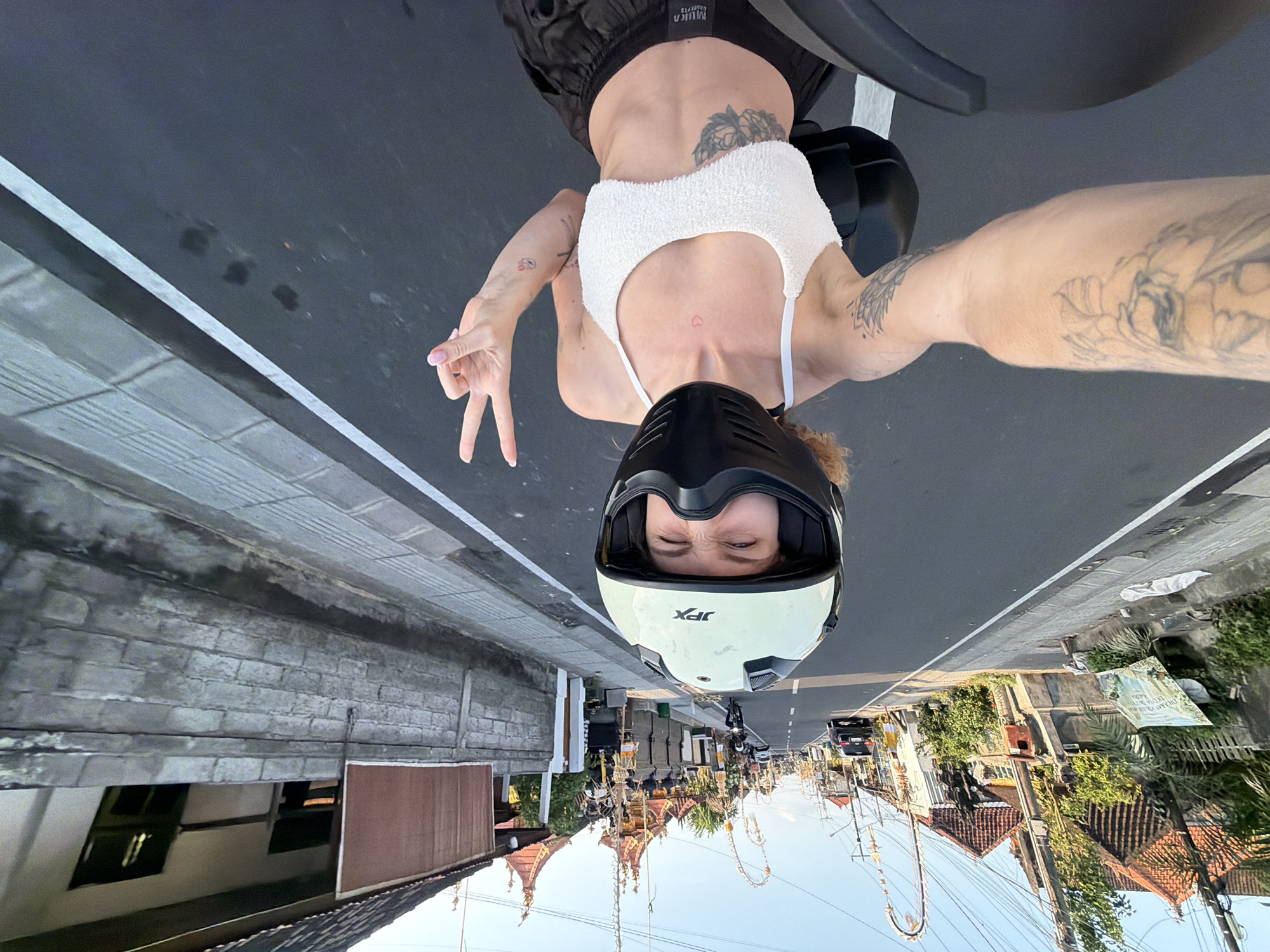 A woman wearing a white helmet and a white crop top is taking a selfie while riding a motorcycle down a street in Mengwi, Indonesia. She is making a peace sign with her right hand. Tattoos are visible on her left arm and abdomen. The street is paved and has a white line marking the lane. Buildings with traditional Balinese architecture line the street, adorned with decorative banners and flags. Power lines crisscross above. A car and other motorcycles are visible in the background. The sky is clear and blue.