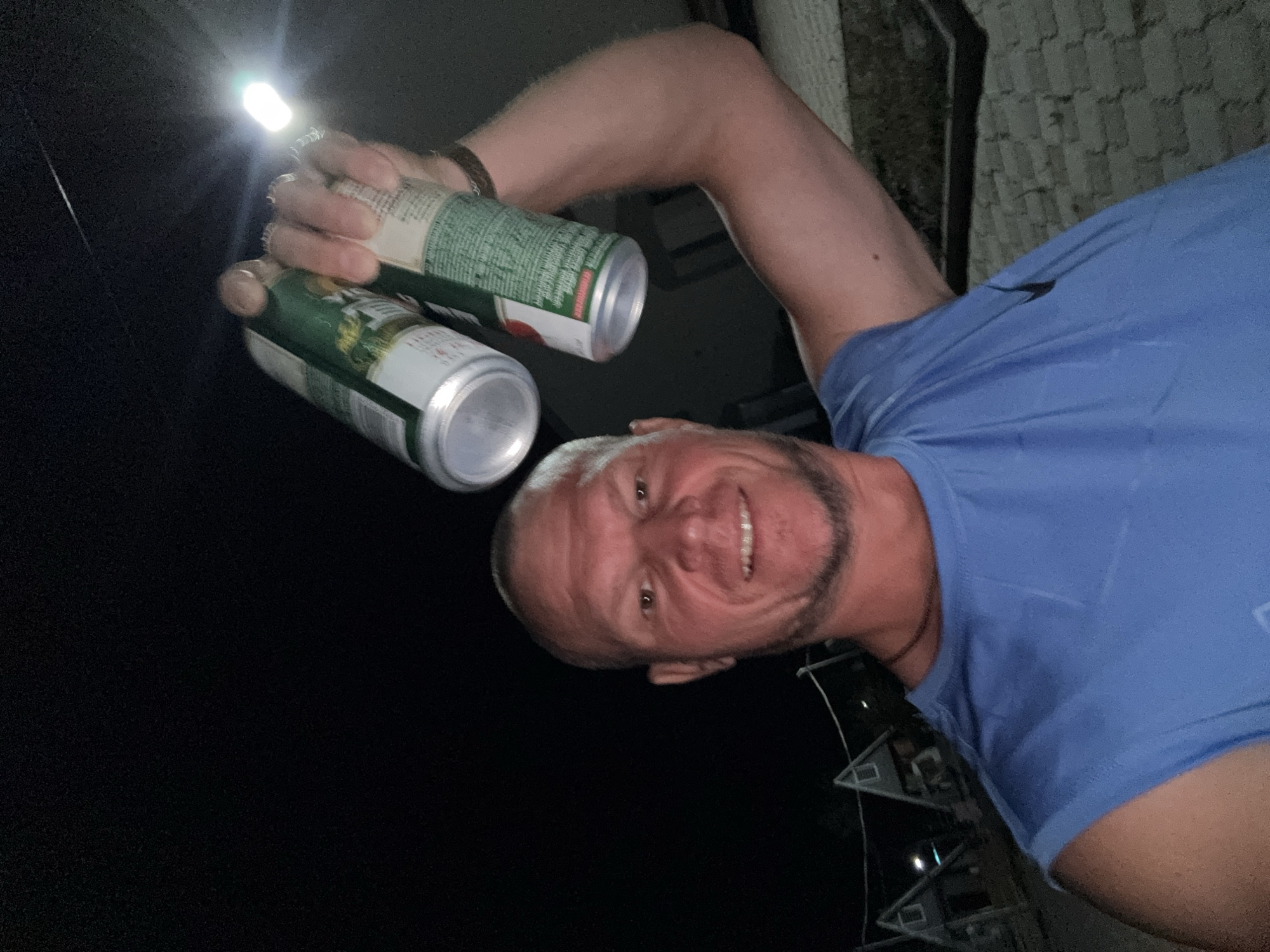 A man is taking a selfie at night in what appears to be an outdoor, possibly rural, setting. He is wearing a blue t-shirt and holding up two aluminum cans. The man has a shaved head, a beard, and is smiling. In the background, there are several triangular-shaped structures that could be houses or cabins, and the ground appears to be paved with bricks. The sky is dark and there is a bright light source in the upper right quadrant of the image, possibly a flashlight or street light. It is not possible to determine the exact location within the city of Urdzhar, Kazakhstan from the visual information provided in the image.