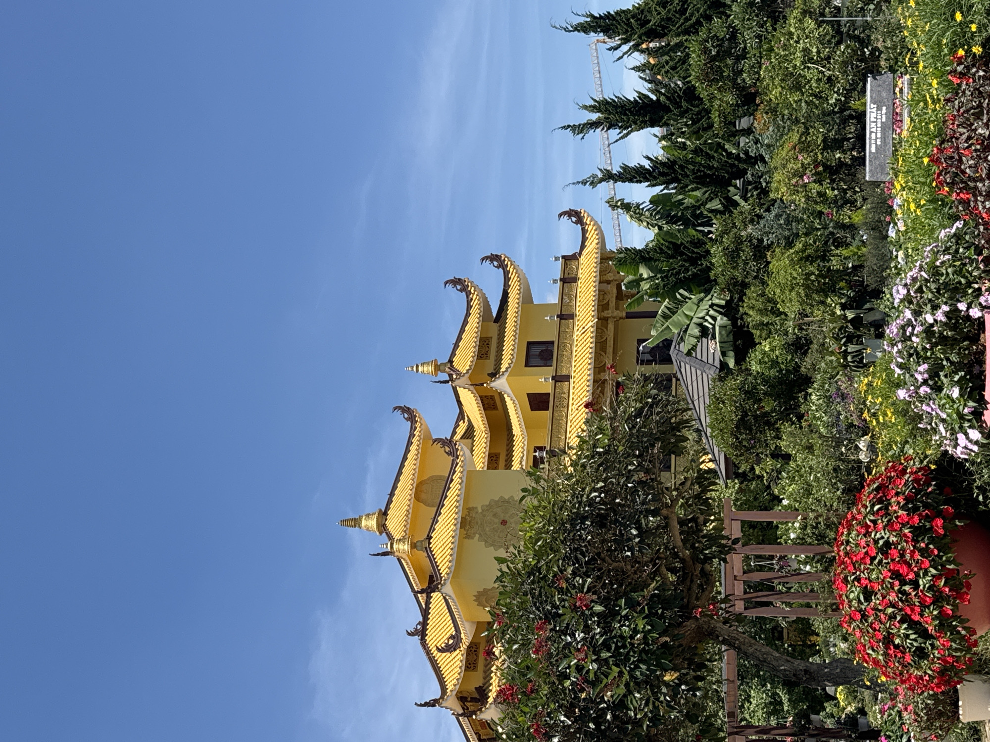The image captures a golden pagoda in Da Lat, Vietnam, set against a clear blue sky. The pagoda has multiple tiers with curved, ornamented roofs and a light yellow facade. Lush greenery surrounds the pagoda, including trees, flowers, and garden arrangements with red blooms. A wooden fence is partially visible in the foreground. In the distance, the blurred form of a construction crane is visible behind the trees. There are no people visible in the image. A sign with writing in Vietnamese can be seen obscured within the garden.