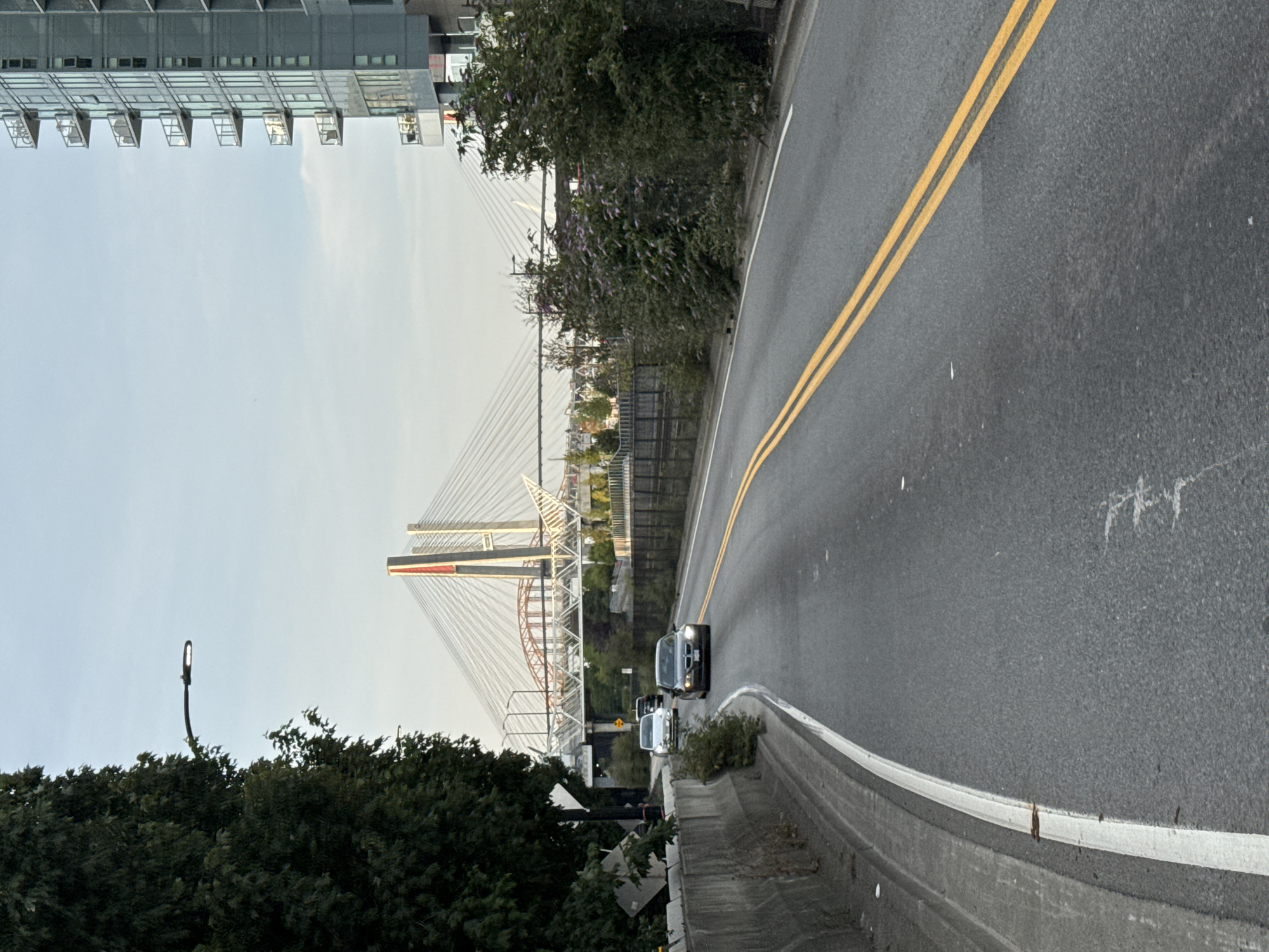 An eye-level shot depicts a street scene in New Westminster, Canada, with a suspension bridge in the background. In the foreground, a grey asphalt road curves to the left, marked with a white line on the left edge and double yellow lines in the center. On the left side of the road, a concrete barrier lines the shoulder, with a large tree growing next to it. A street lamp is visible above the tree. Further down the road, three cars are driving in the same direction. The nearest car is a dark sedan. To the right of the road, a modern, multi-story building with balconies stands tall, its windows reflecting the sky. Beyond the road, a suspension bridge with prominent cables and red and white towers spans the horizon. The sky is overcast with scattered clouds.