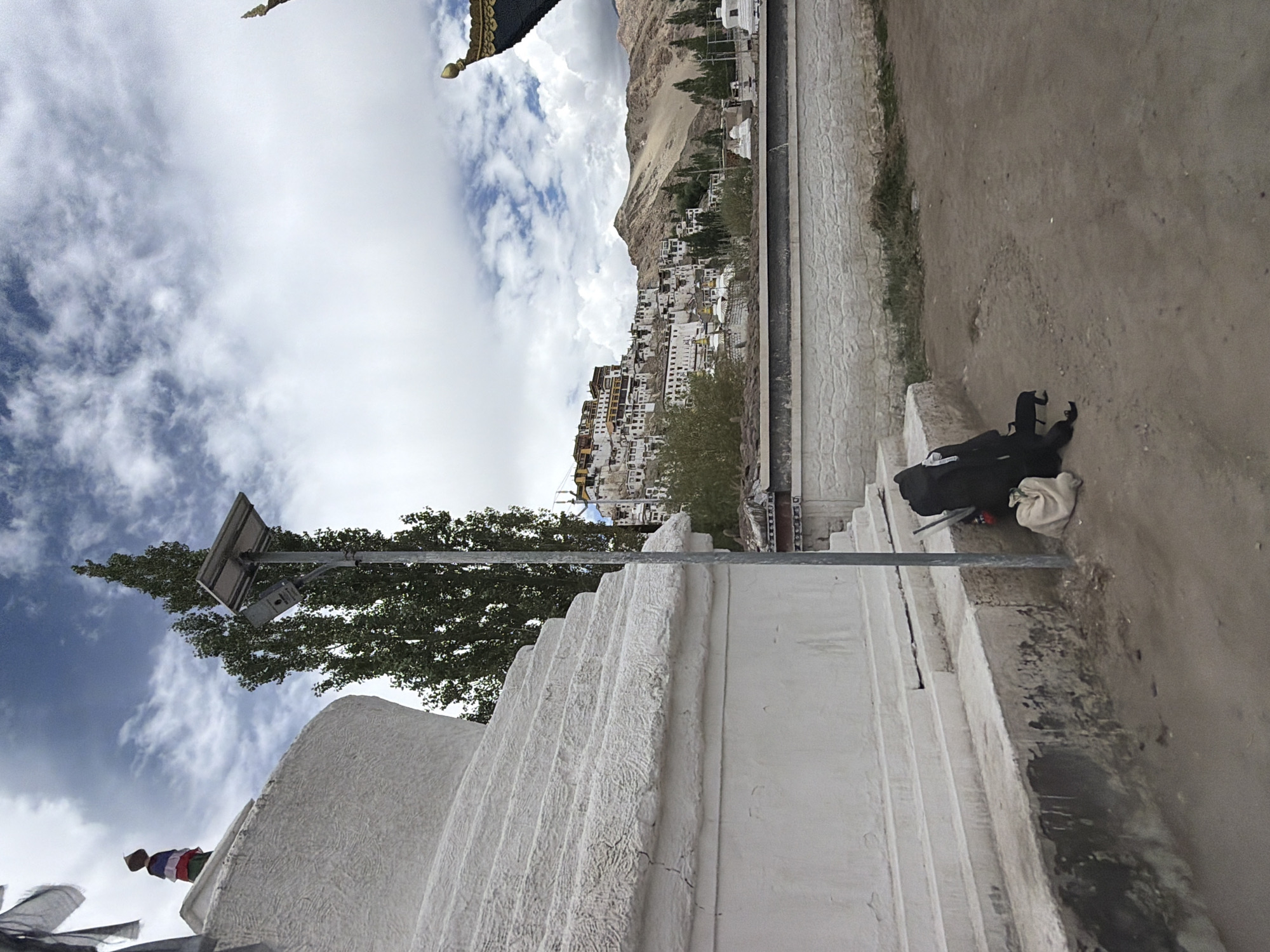 A landscape view of Thiksey Monastery in Ladakh, India, features a large white stupa in the foreground to the left. A tall, slender tree stands behind the stupa, accompanied by a solar-powered street light. To the right of the stupa and street light, a black backpack and a light-colored bag are placed on the ground. In the background, the monastery complex, with its distinctive white and yellow buildings, is visible cascading down a hillside. The sky is filled with dramatic clouds, and the terrain is arid and mountainous.