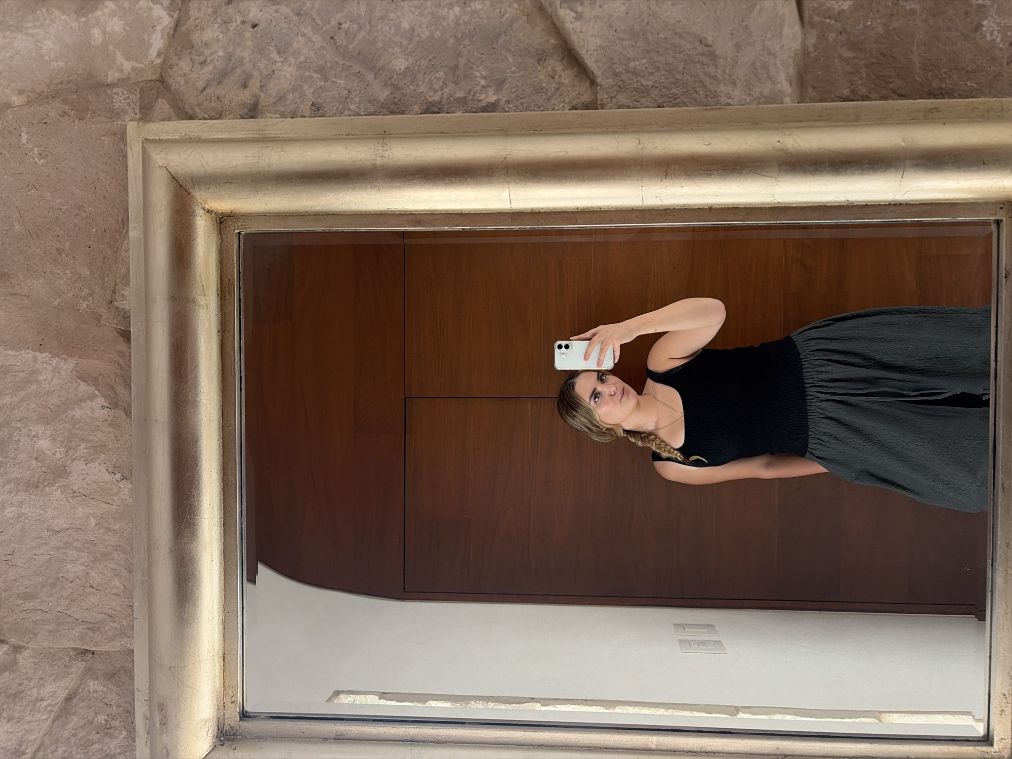 A young woman is depicted in a mirror selfie, holding a white smartphone to capture her reflection. She looks directly at the camera with a neutral to slightly pensive expression. Her blonde hair is styled in a braid over her left shoulder, and she is adorned with a gold necklace and a thin bracelet on her right wrist. She wears a black ribbed tank top and a dark green or grey pleated skirt.
The mirror she is using is framed by a wide, ornate golden border that appears to have a slightly distressed or aged finish. Outside the mirror's frame, primarily visible on the left and top edges, is a textured wall constructed from large, light brown or beige stone blocks, suggesting a rustic or traditional architectural setting.
Reflected within the mirror, behind the woman, is a prominent dark brown wooden wall or large wooden door. Below this wooden section, a segment of a white wall is visible, featuring a rectangular electrical outlet or light switch. The overall scene suggests an indoor setting, likely a room within a residence or building. There is no visible text in the image. The location context provided is San Bernardino Tlaxcalancingo, Mexico.