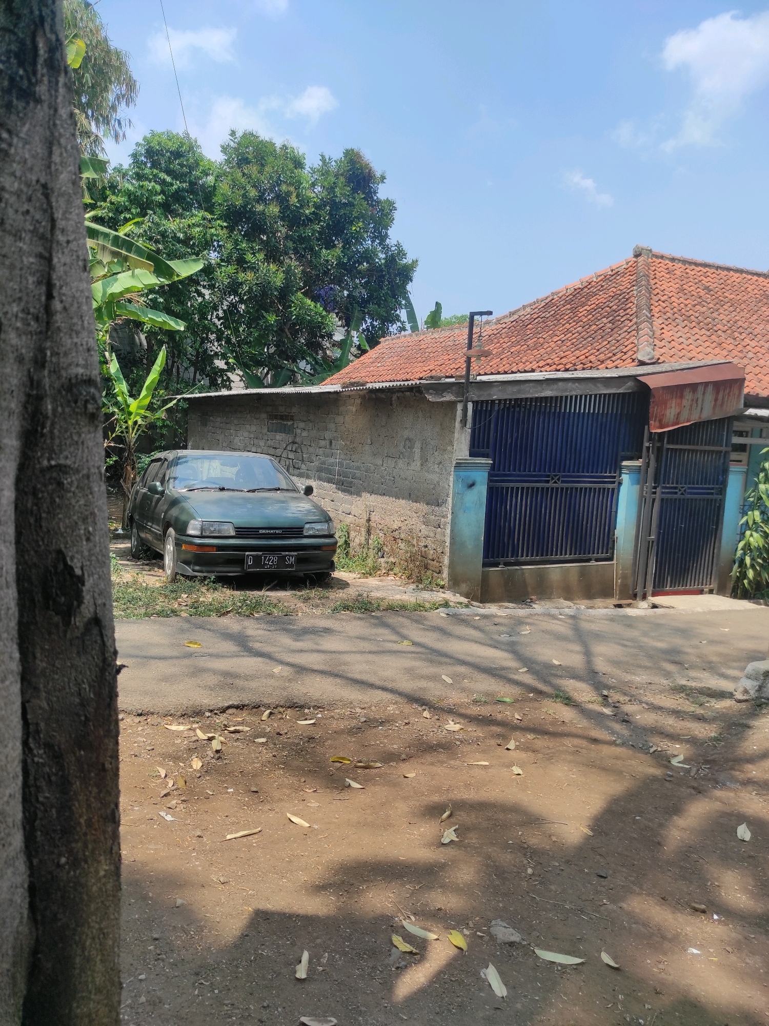 This is a vertical, eye-level, medium shot of a street scene in Padalarang, Indonesia, on a sunny day. In the foreground, a large tree trunk on the left partially obscures the view. The paved street in front of the tree is sparsely littered with dry leaves and shadowed by dappled sunlight.
The mid-ground features a green, four-door sedan parked on the side of the street, facing the viewer. The car has a license plate that reads "D 1428 SM". Behind the car, a rough, light-colored brick wall extends across the scene. To the right of the wall, a house with a distinctive orange, tiled roof is visible. The house has a dark blue metal gate and fence, with some lighter blue trim around the entrance. Green foliage, including banana leaves, grows around the house and car.
The background is dominated by a bright blue sky with scattered white clouds and the dense green canopy of trees. The overall impression is a residential area with a car parked outside a house, under a clear sky.