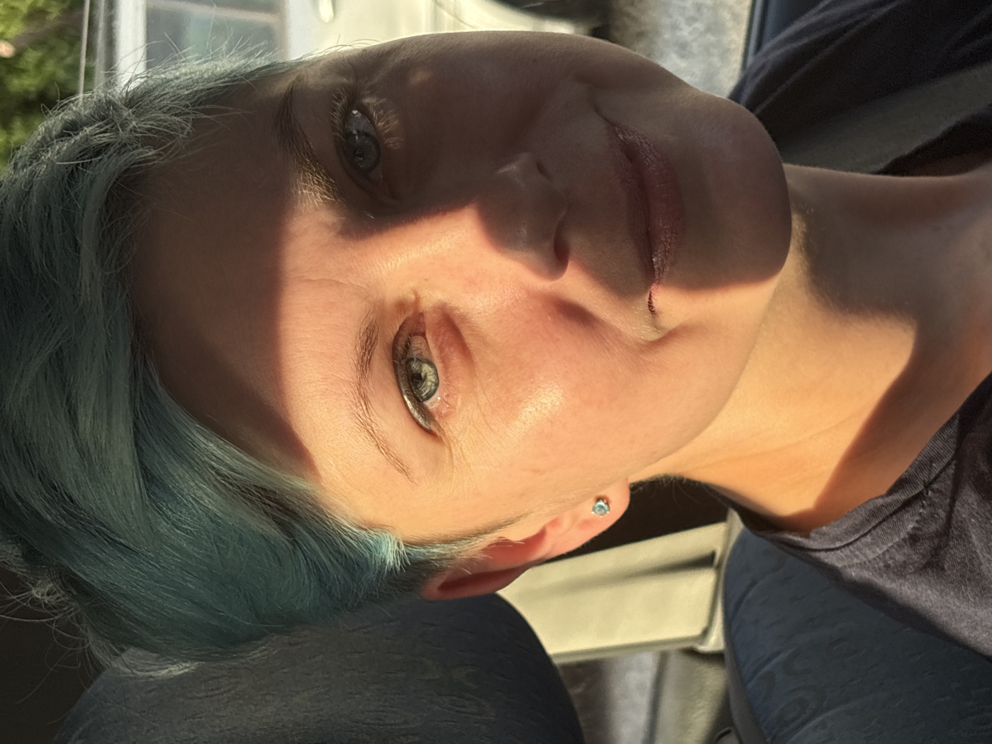 A close-up, rotated portrait captures a person with distinctive short, teal-colored hair and light blue-green eyes. The subject, likely female, has a fair complexion and is looking towards the camera with a gentle, relaxed smile. A significant portion of the face is illuminated by strong, direct sunlight, creating a dramatic shadow that diagonally crosses the nose and mouth, leaving the left side of the face in partial shadow. A small, light-colored stud earring is visible in the right earlobe.
The subject is wearing a dark, possibly navy or black, casual top, with a glimpse of fabric visible around the neck and shoulder area. The background is softly blurred but suggests an indoor setting, possibly within a vehicle given the soft textures and hints of what might be a seat or interior panel. The lighting indicates it is daytime, likely morning or late afternoon, when the sun is lower in the sky, casting long shadows. No specific location cues are visible in the image itself, but the provided context places it in Bar, Montenegro. There is no visible text. The overall impression is a casual, sunlit moment.
